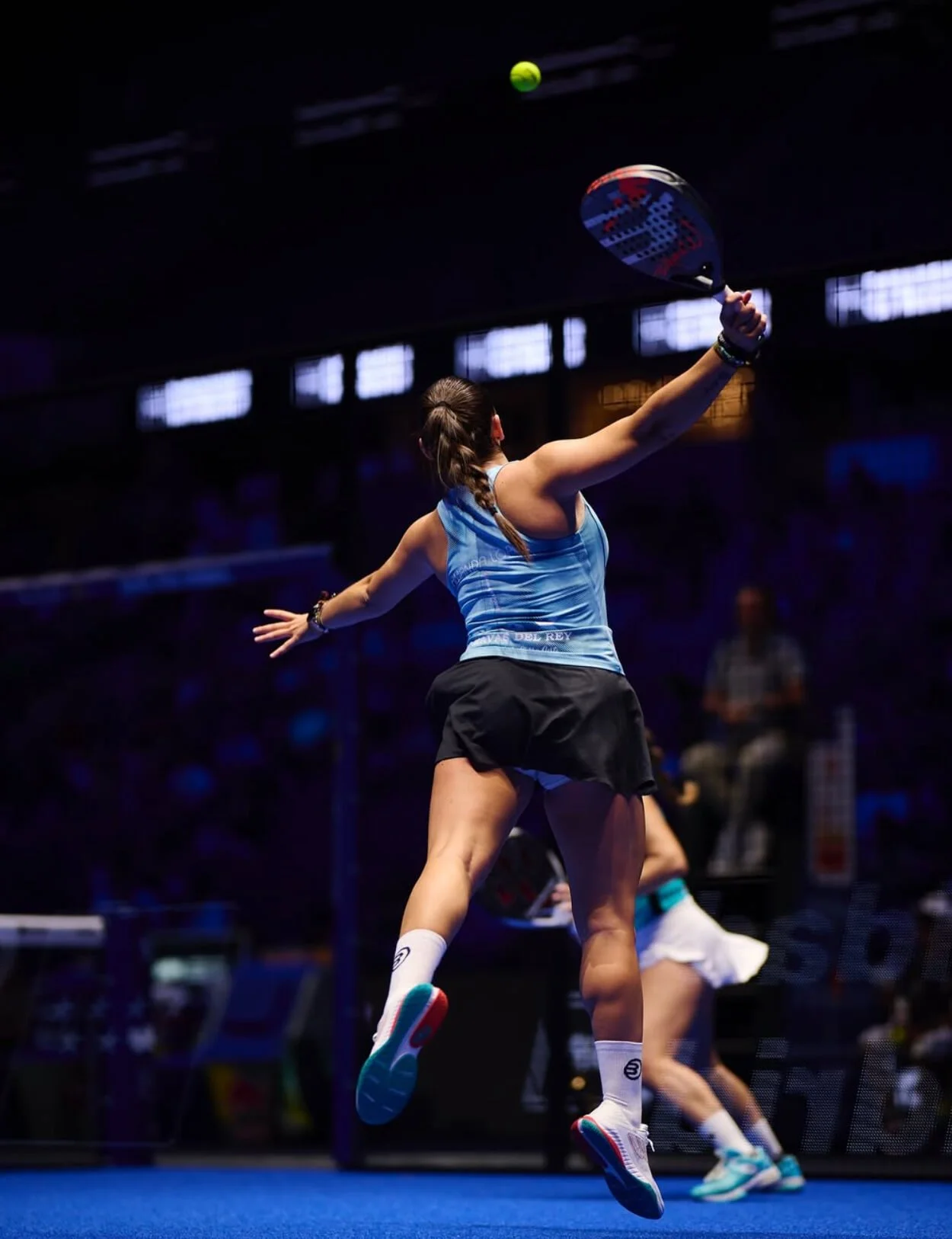 A female padel player in a blue top and black skirt hitting a padel ball during a match indoors, with other players and officials visible in the background.