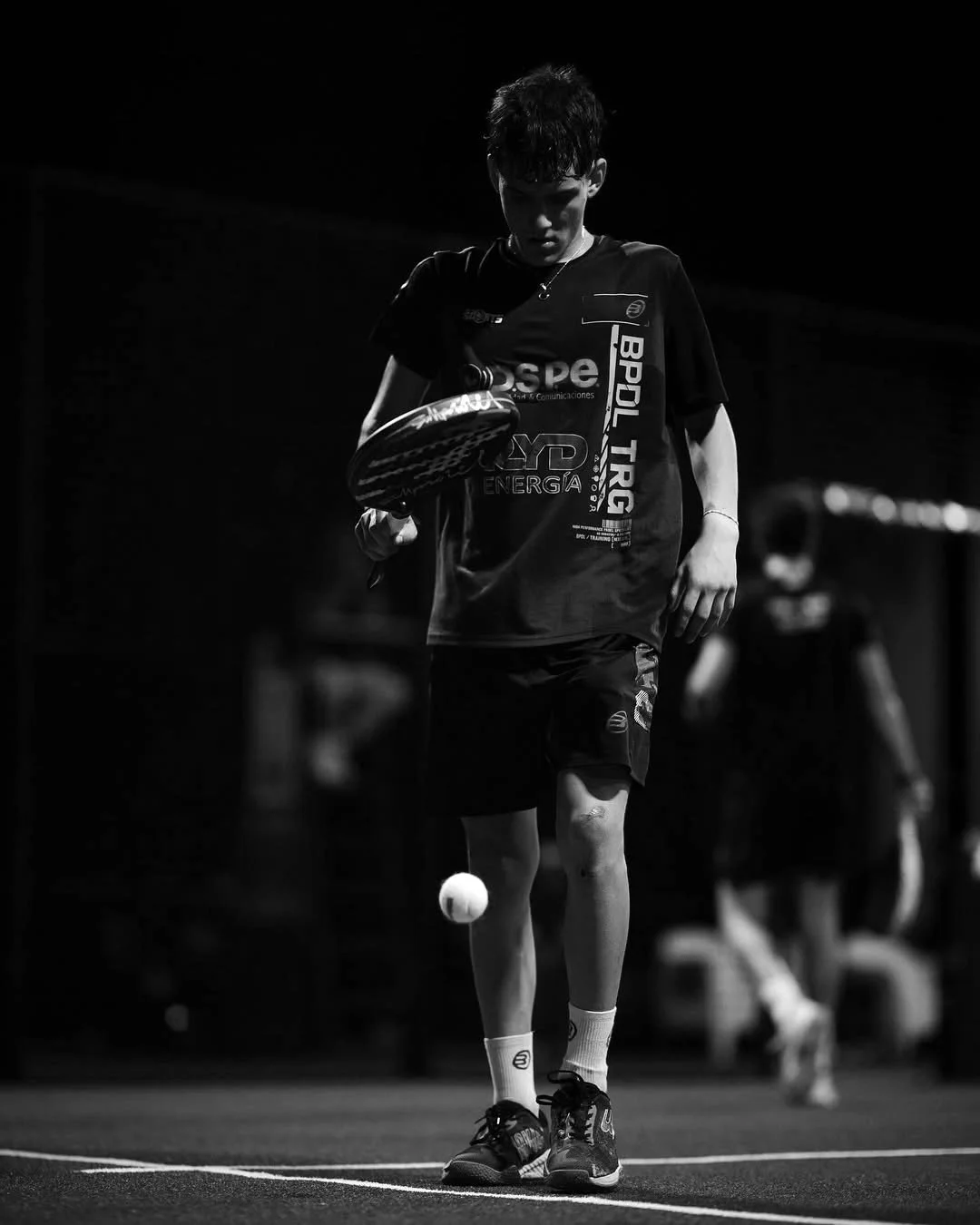 A young padel player is on a court, preparing to hit a ball with a paddle. He is dressed in sportswear and focused on the game, with a dark background and other players faintly visible in the distance.