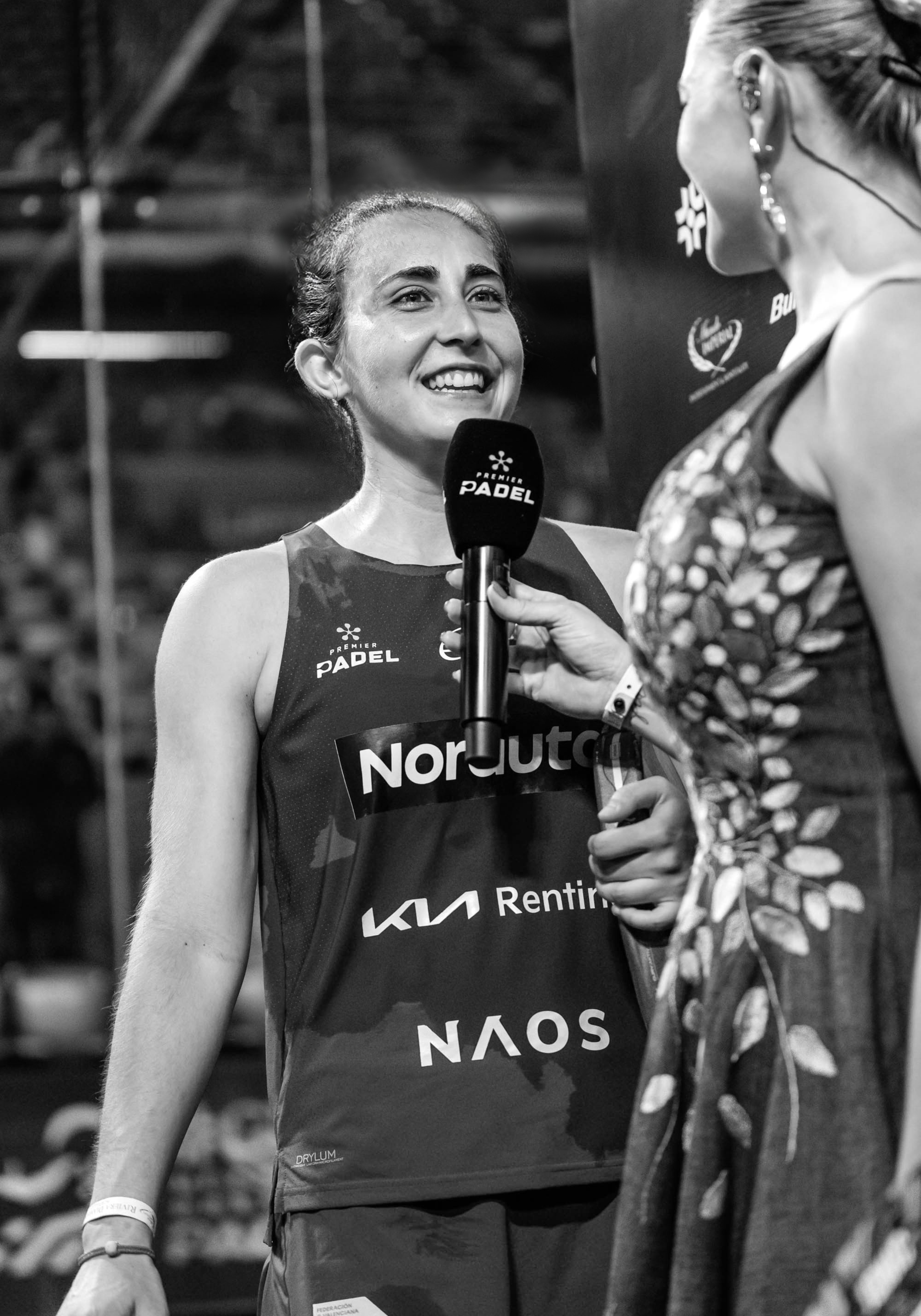 A female padel player with a big smile holding a microphone during an interview, wearing a sports jersey with sponsor logos, in an indoor sports venue.