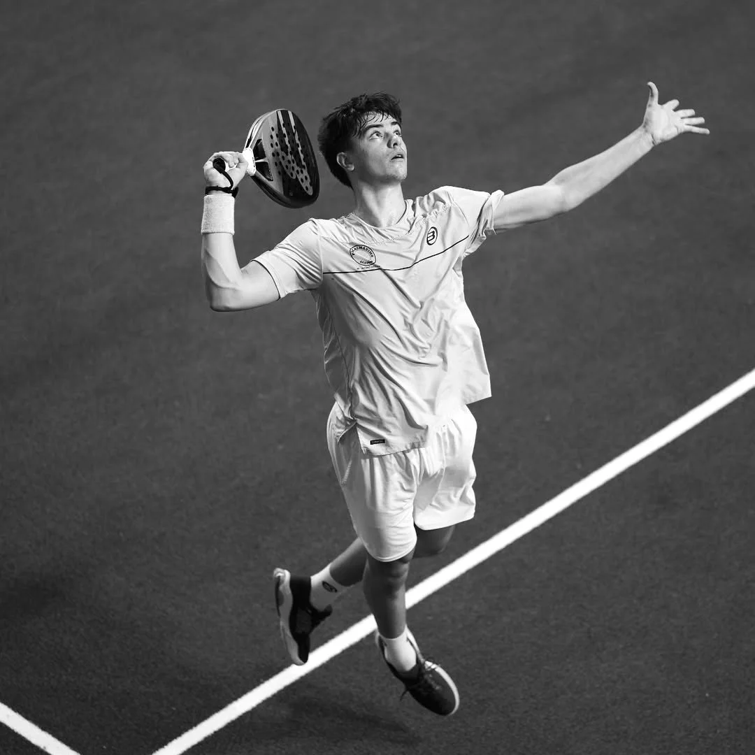A male padel player is in mid-air, preparing to hit a serve on a padel court. He is holding a padel racket in his right hand, wearing a sport shirt, shorts, and sneakers, and is extending his left arm outward.