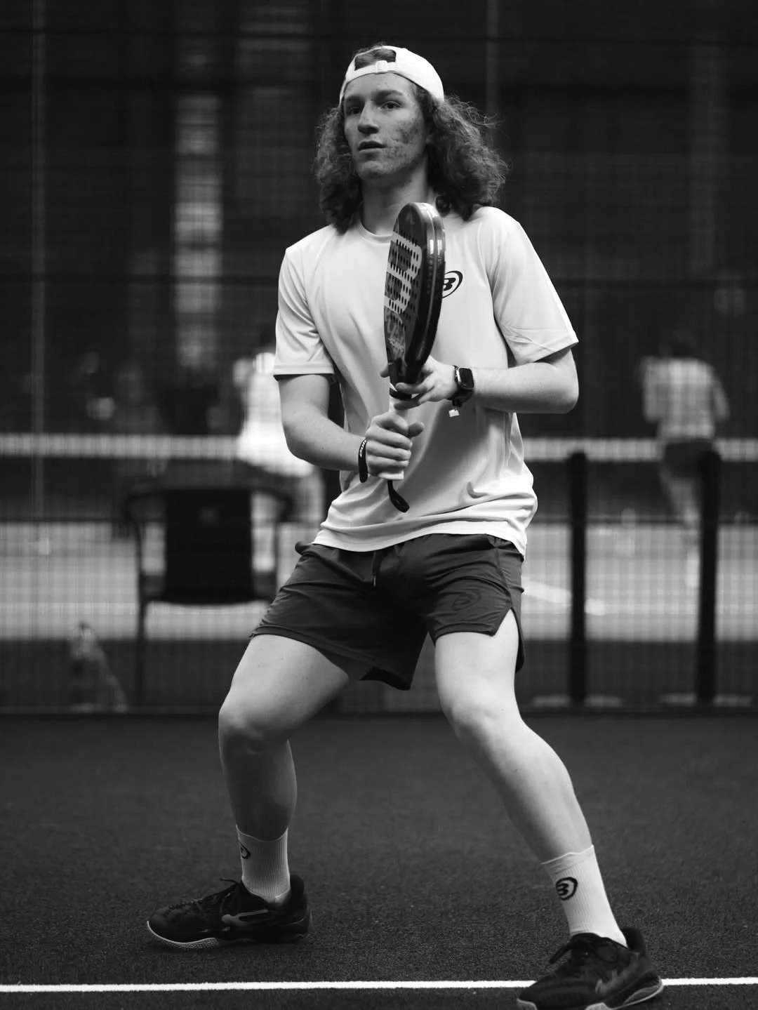 A young man with curly hair, wearing a baseball cap backwards, a t-shirt, shorts, and athletic shoes, holds a tennis racket and prepares to hit a ball during a padel match on an indoor court.