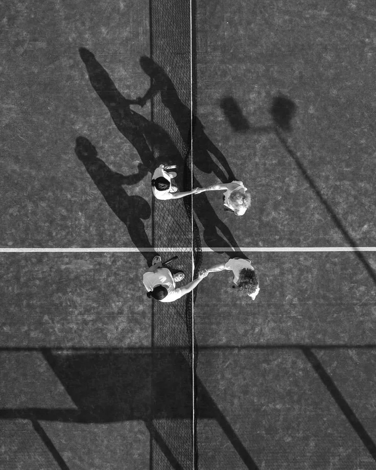Top-down black and white photo of two tennis players shaking hands at the net after a match on an outdoor court, with their shadows cast on the court surface.