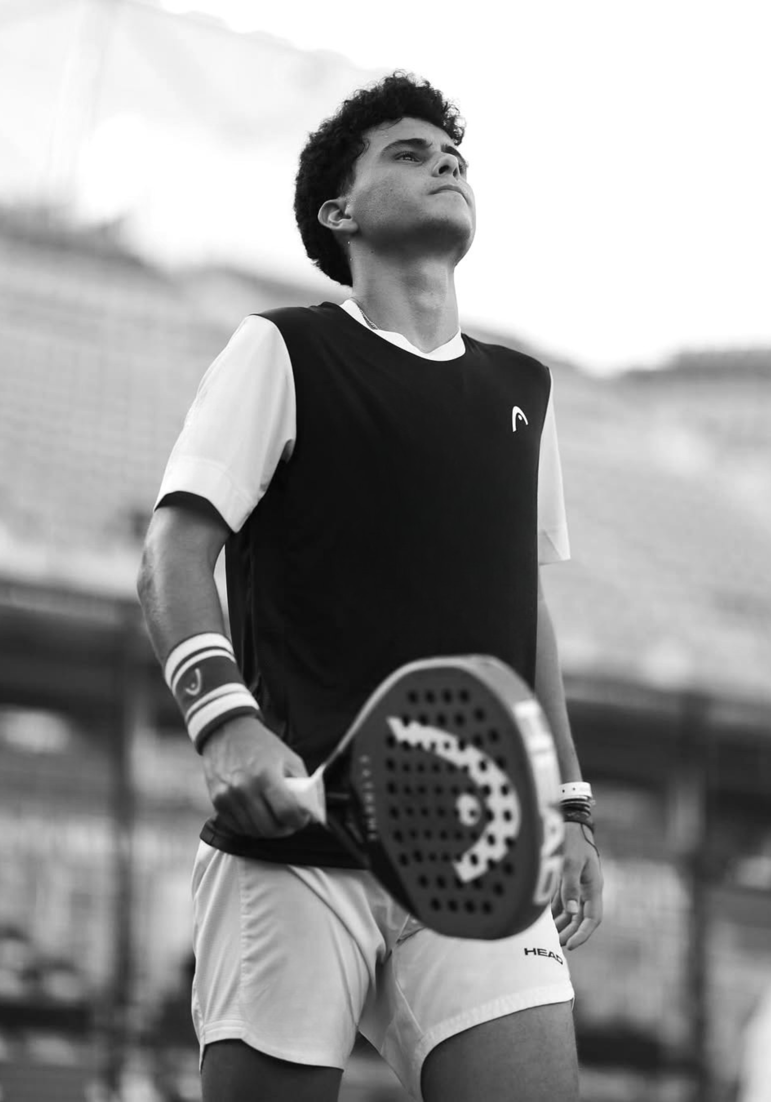 Black and white photo of a male padel player holding a tennis racket on a tennis court, looking upward with a determined expression.