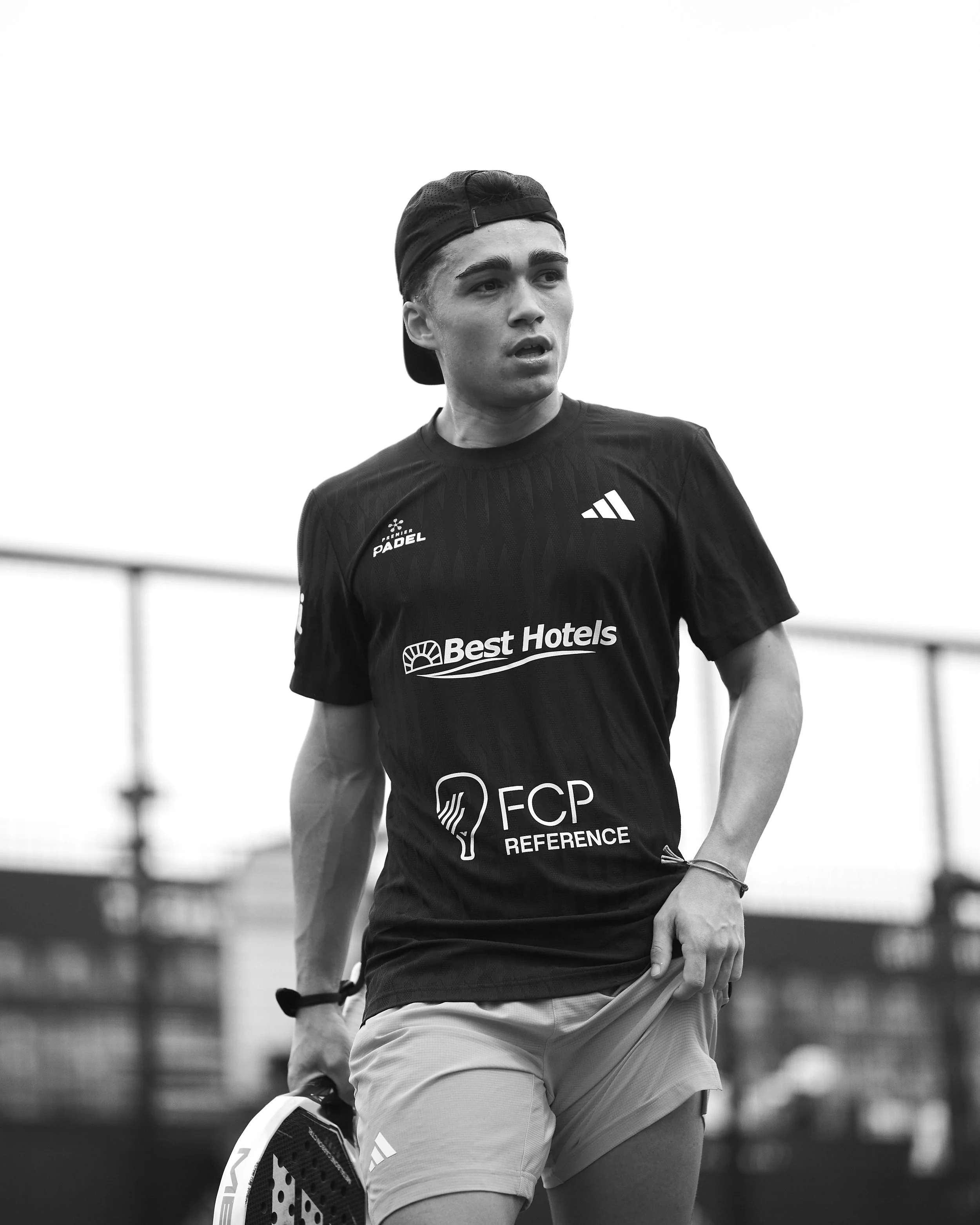 A young male athlete in sports attire, wearing a black sports T-shirt with logos and text, and a backward cap, standing outdoors holding a padel racket in his left hand. The background is blurred with a fence and some structures visible.