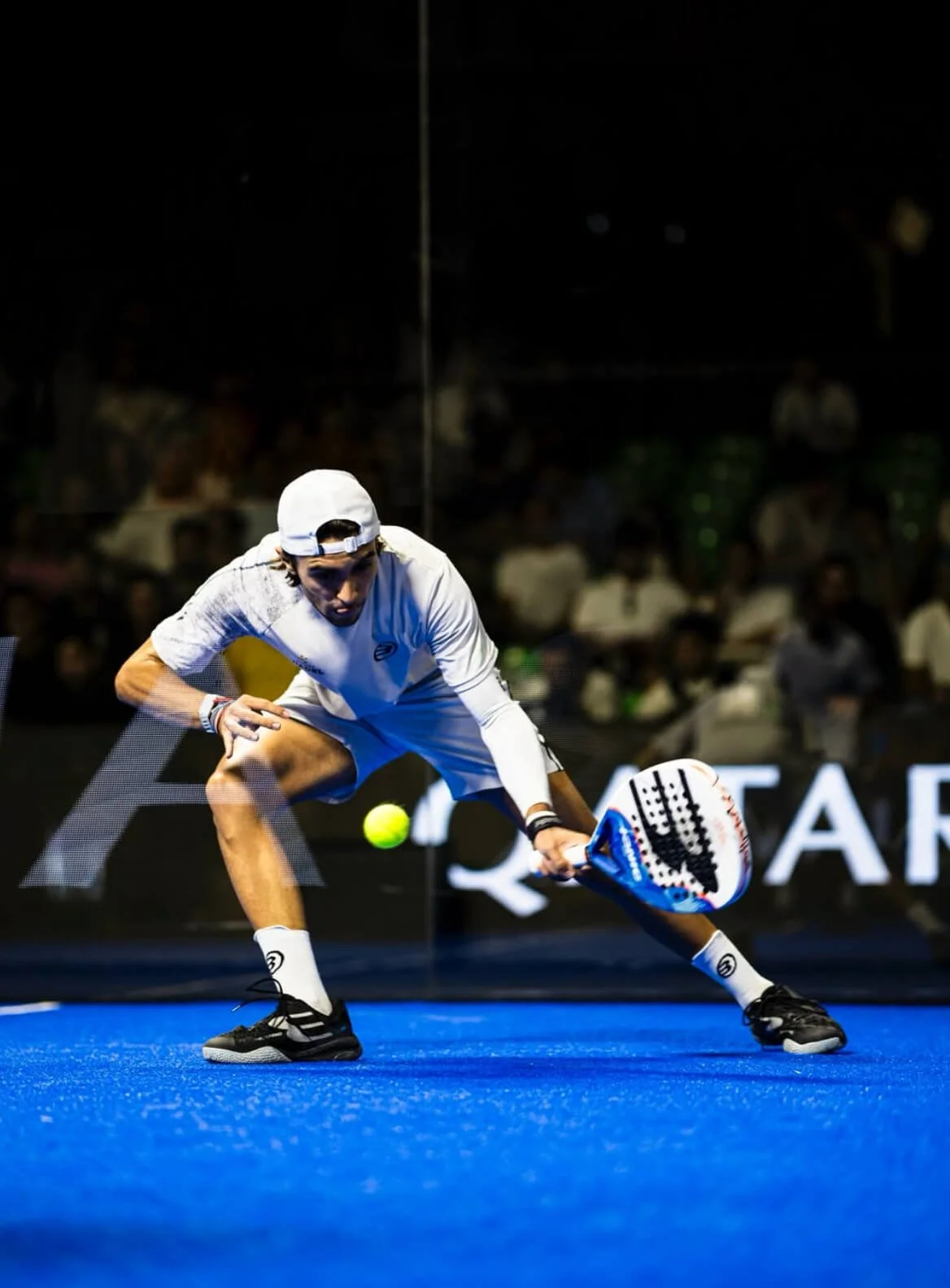 A padel player in white athletic wear and a backward cap reaching to hit a tennis ball with his racket on a blue court during a match.