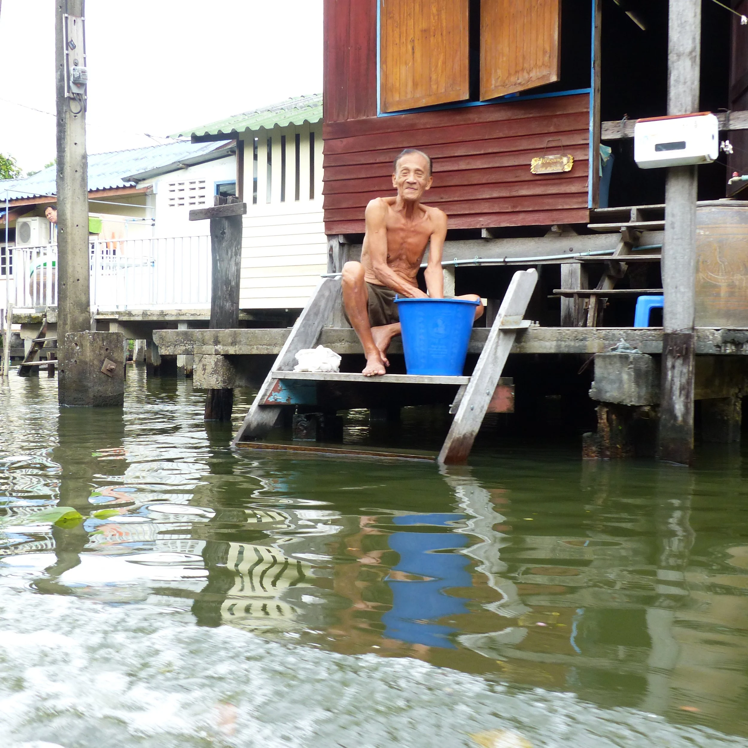 Un homme adulte assis en plein air près d'une maison en bois, avec de l'eau en dessous et un seau bleu devant lui. La maison est en bois, sur pilotis, avec une petite escarre en bois. En arrière-plan, il y a une autre maison en couleur claire avec un balcon, et une personne regarde vers l'objectif.