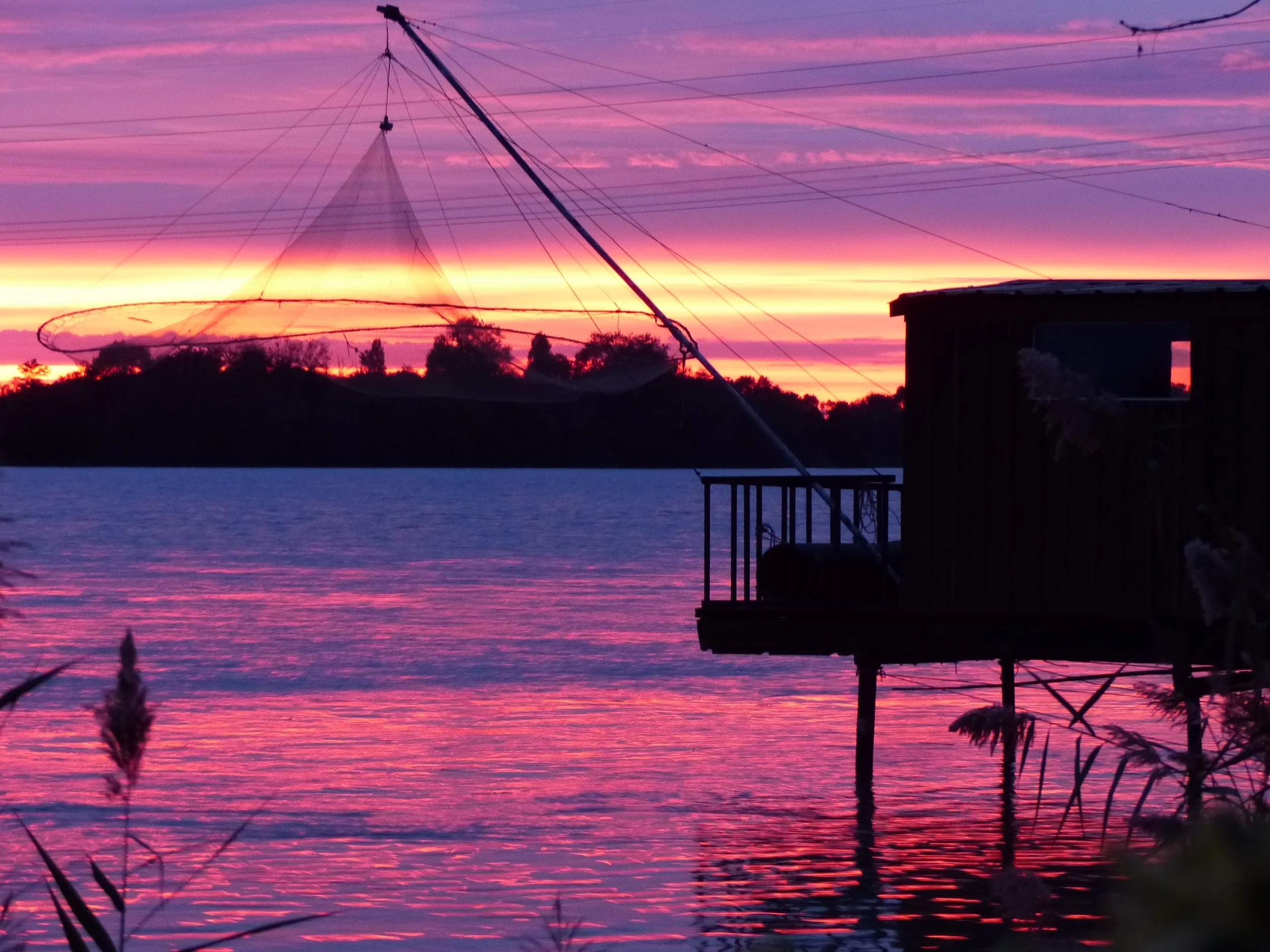 Coucher de soleil sur un lac avec un ciel coloré en nuances de violet, rose et orange, et des silhouettes de vegetation et d'une cabane en bois en premier plan.