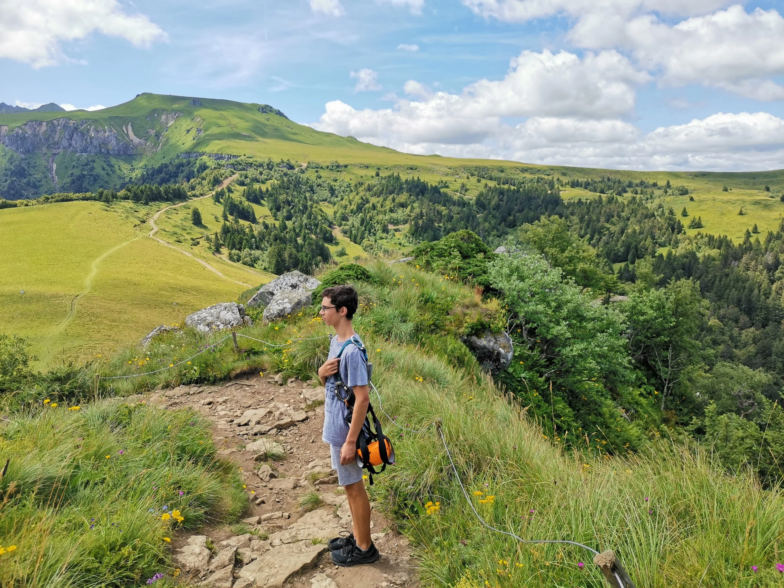 Jeune garçon portant un T-shirt gris et un sac à dos orange, observant un sentier de randonnée en montagne avec des collines verdoyantes, des arbres et un ciel partiellement nuageux.