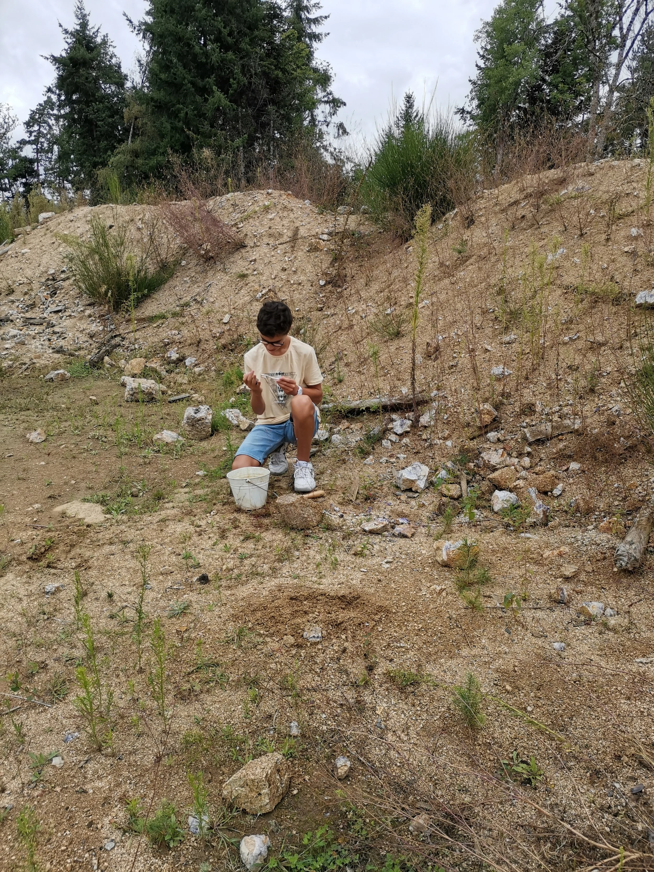 un jeune garçon assis sur le sol, regardant un objet dans ses mains, avec un seau blanc à côté, dans un environnement rocheux avec des herbes et des arbres en arrière-plan.