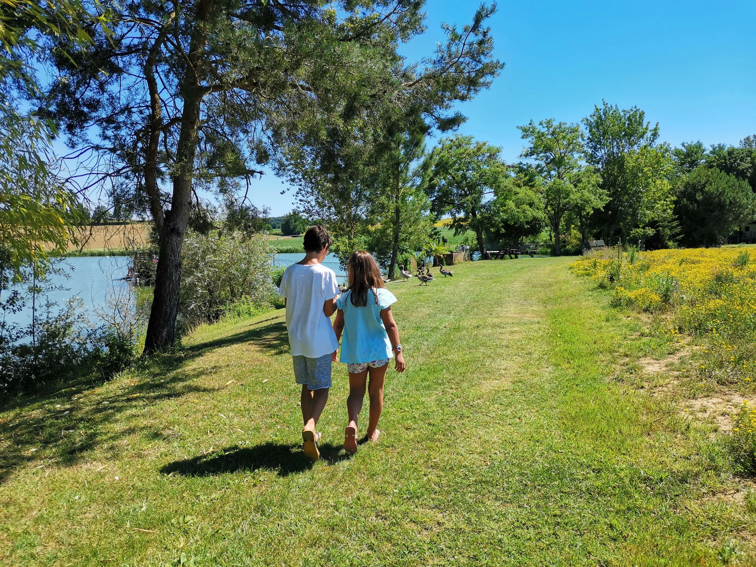 Deux enfants marchent dans un parc verdoyant près d'un étang, entourés d'arbres et de fleurs, par une belle journée ensoleillée.