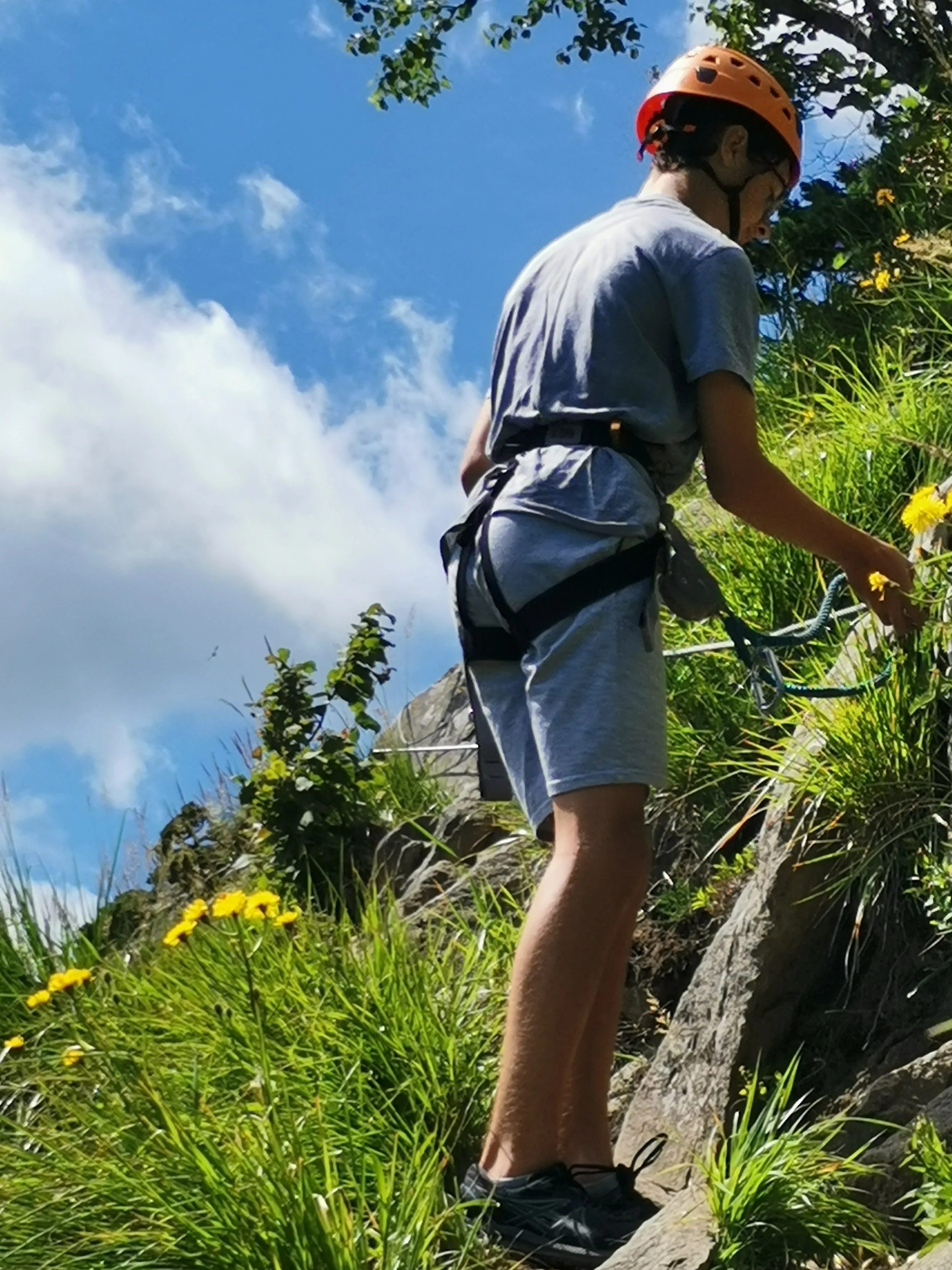 Un jeune homme avec un casque orange grimpe une falaise rocheuse entouré de végétation verte et de fleurs jaunes, sous un ciel bleu avec quelques nuages.