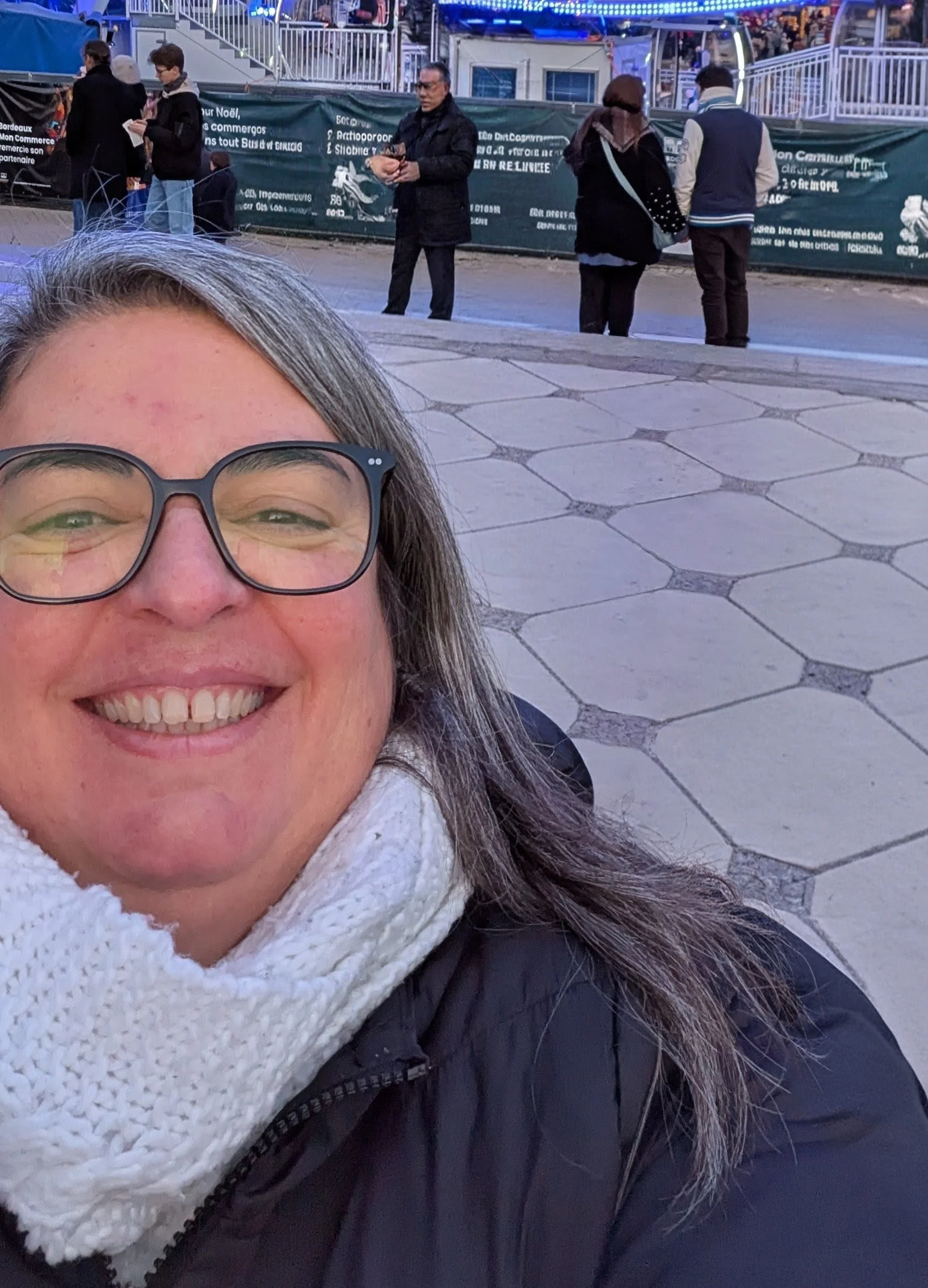 Une femme souriante portant des lunettes, avec un foulard blanc, une veste noire et des cheveux gris. En arrière-plan, il y a plusieurs personnes qui se tiennent derrière une barrière, et un manège avec des lumières bleues.