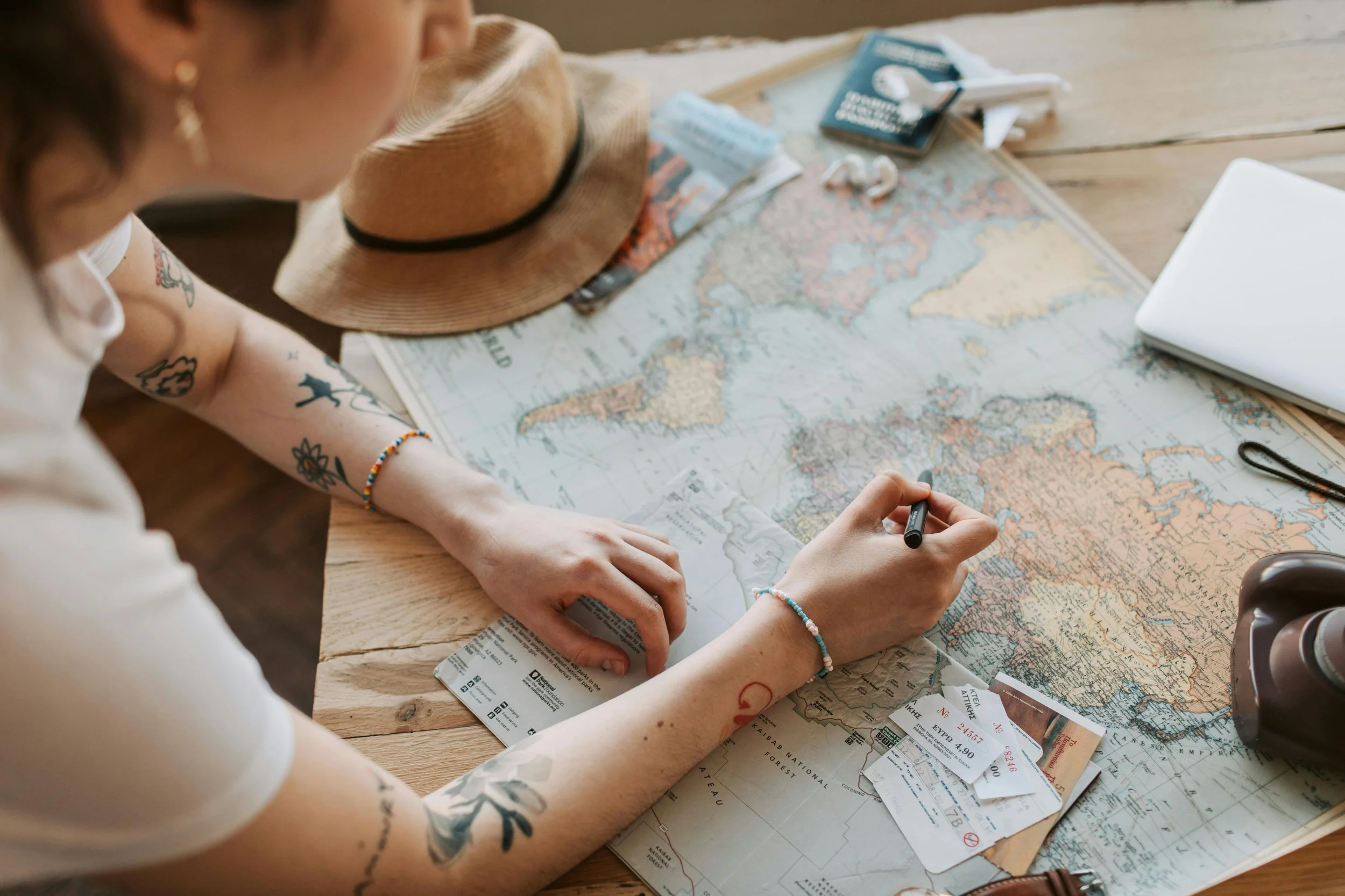 Une personne regarde une carte du monde sur une table en bois, avec un chapeau, des billets d'avion, un ordinateur portable et des objets de voyage autour.