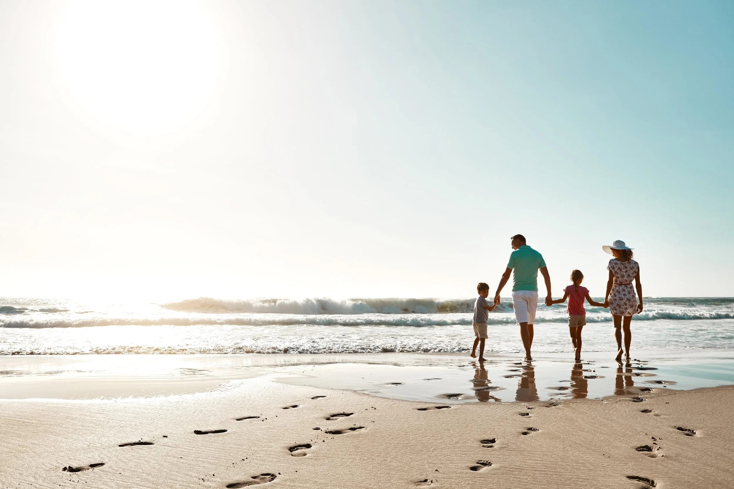 Une famille se promène sur la plage ensoleillée, laissant leurs empreintes dans le sable et tenant la main. La mer et le ciel sont visibles en arrière-plan.
