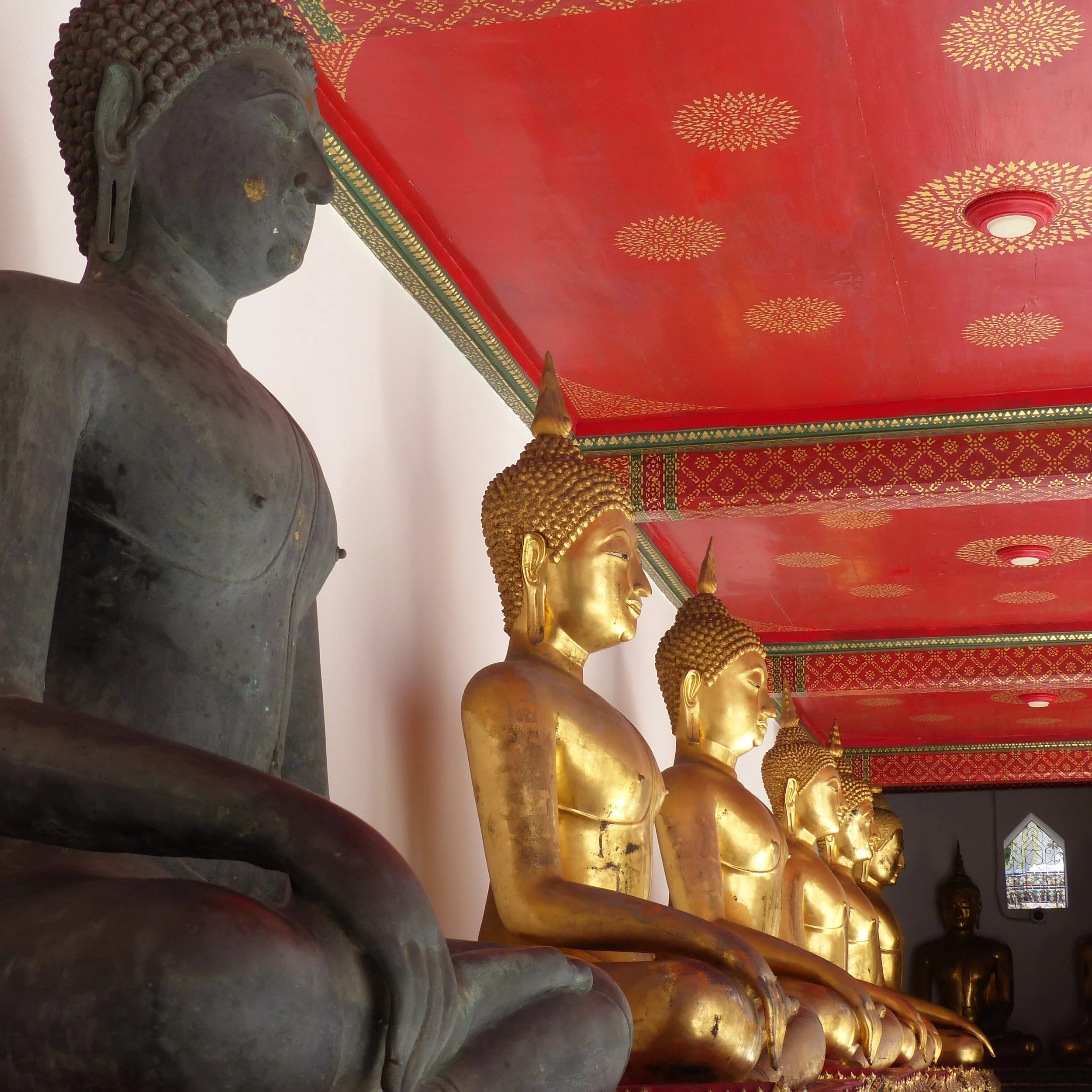 Une rangée de statues dorées de Bouddha alignées en dessous d'un plafond rouge décoré de motifs dorés, avec un plafond en partie visible.