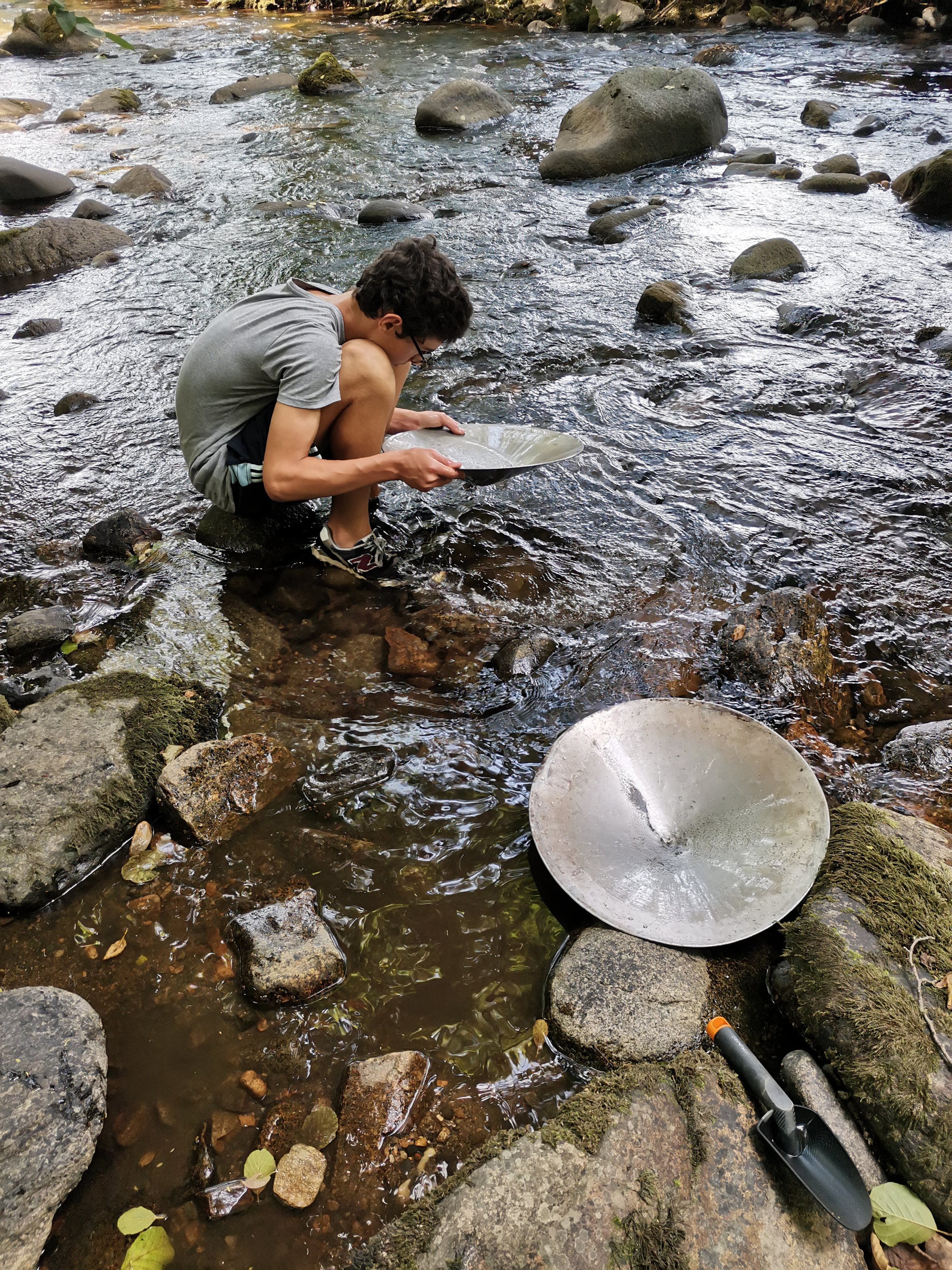 Une personne assise sur un rocher dans une rivière, tenant une cuillère ou un petit plat. Il y a deux surfaces métalliques rondes dans l'eau, probablement des pales de rotor. La rivière est bordée de pierres et de feuilles, l'eau coule doucement.