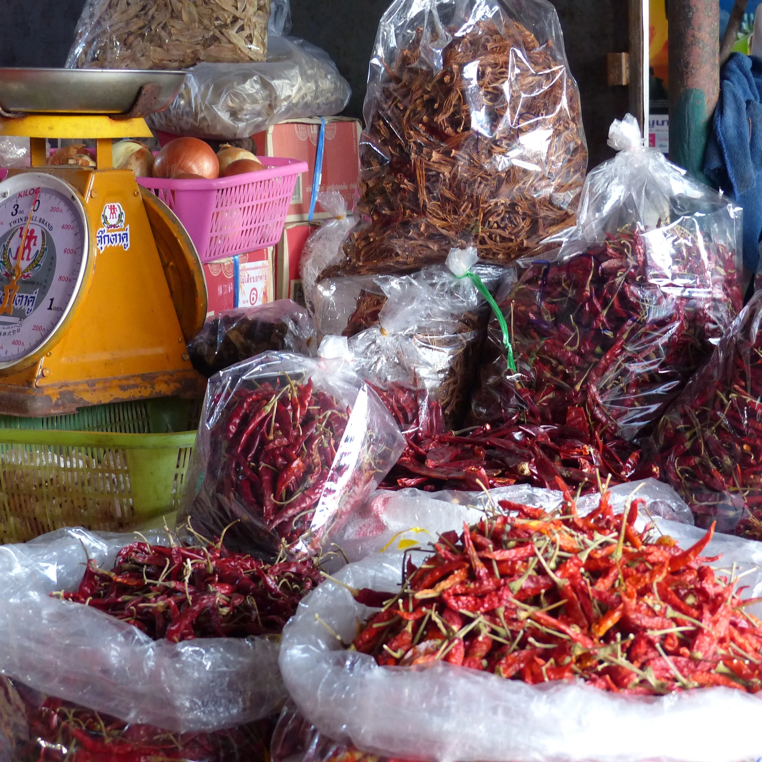 Divers sachets en plastique contenant des piments rouges séchés sur un marché, avec une balance jaune et des oignons en arrière-plan.