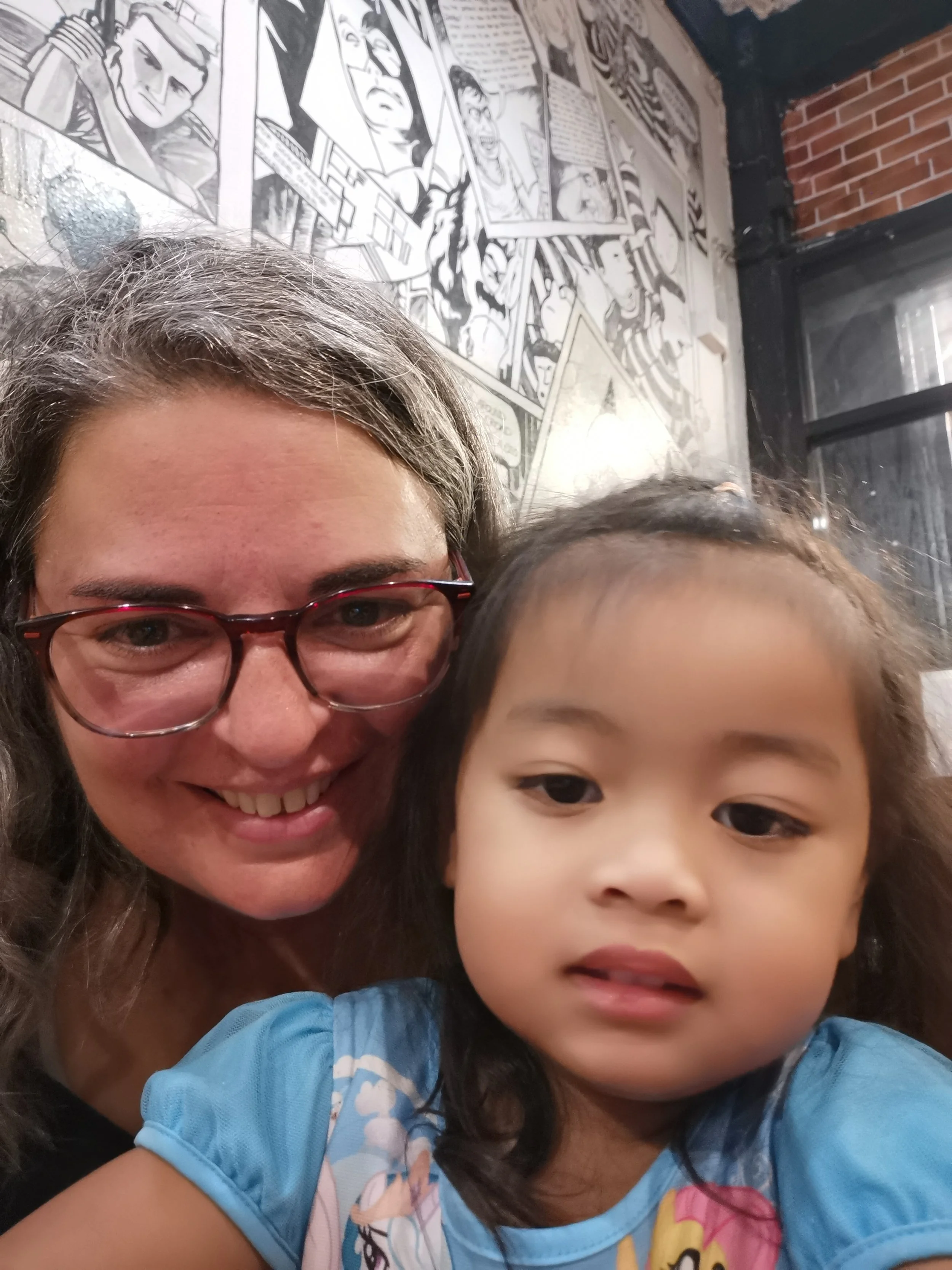 Une femme avec des lunettes et un jeune enfant prennent un selfie ensemble, en arrière-plan un mur avec une œuvre d'art murale en noir et blanc, ainsi qu'un mur de briques et une fenêtre.