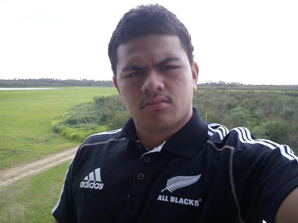 A young man taking a selfie outdoors on a grassy area with a dirt path and green trees in the background, wearing a black rugby shirt with Adidas and All Blacks logos.