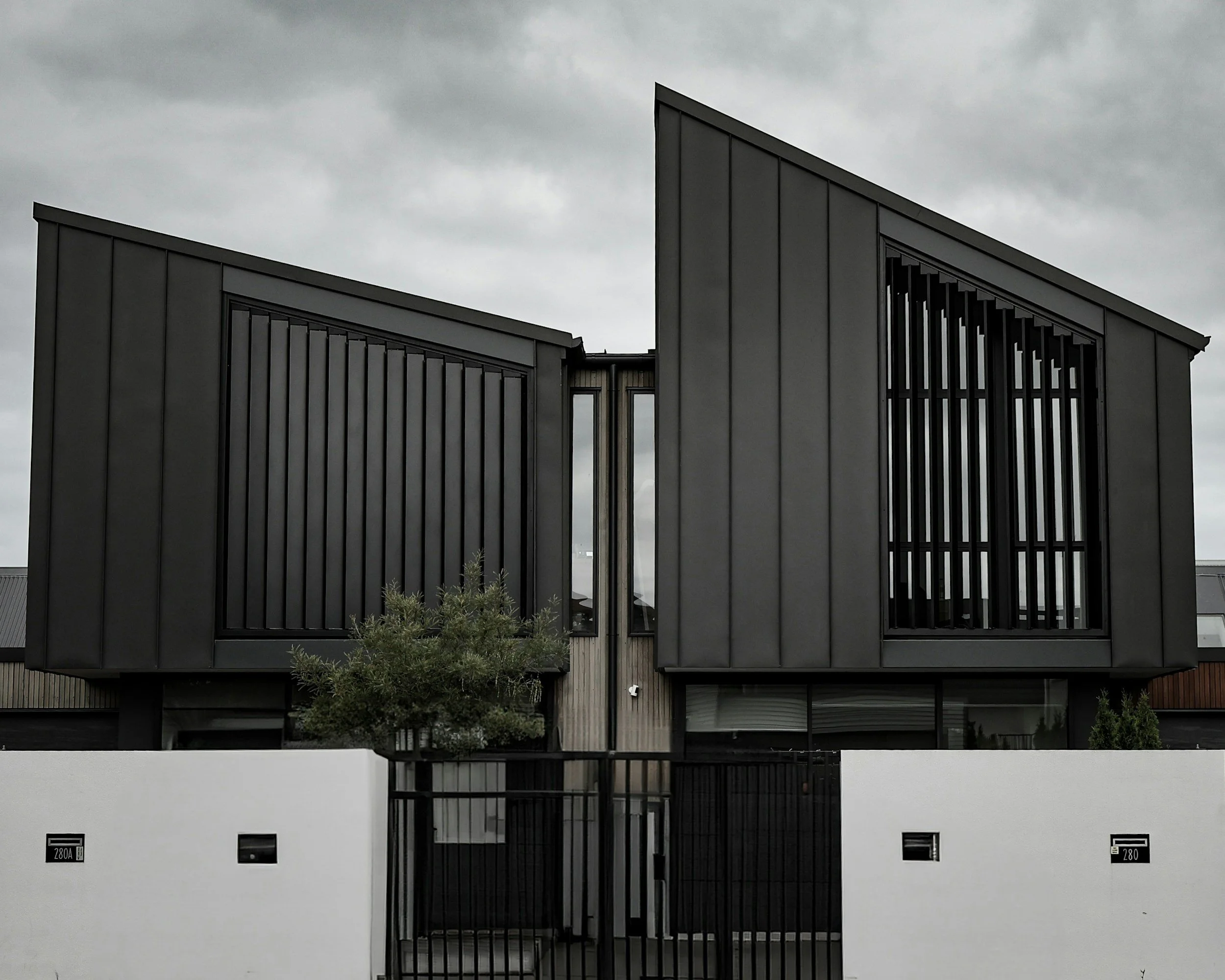 Modern black house with angular design and vertical slats, surrounded by a white fence, overcast sky.