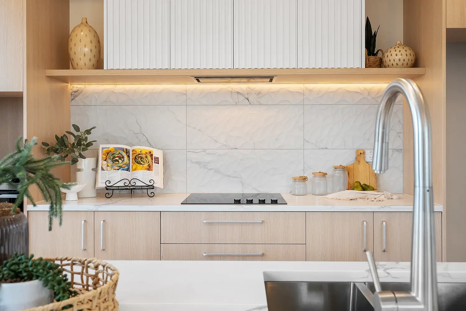 Modern kitchen with light wood cabinetry, white marble backsplash, and a stainless steel faucet. Decorative vases and jars are on the shelves, and a cookbook on a stand is on the countertop.