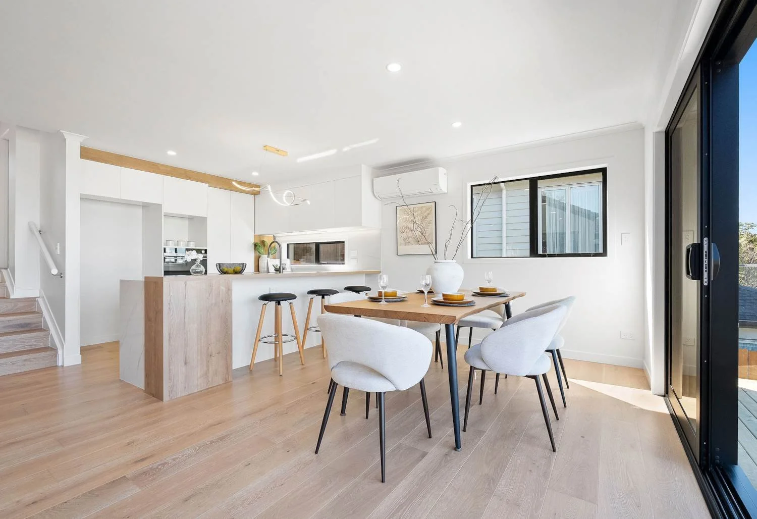 Modern open-plan kitchen and dining area with light wood flooring, white walls, and black-framed windows. The dining table is set with bowls, plates, and wine glasses, surrounded by white upholstered chairs. There are black bar stools at a white kitc