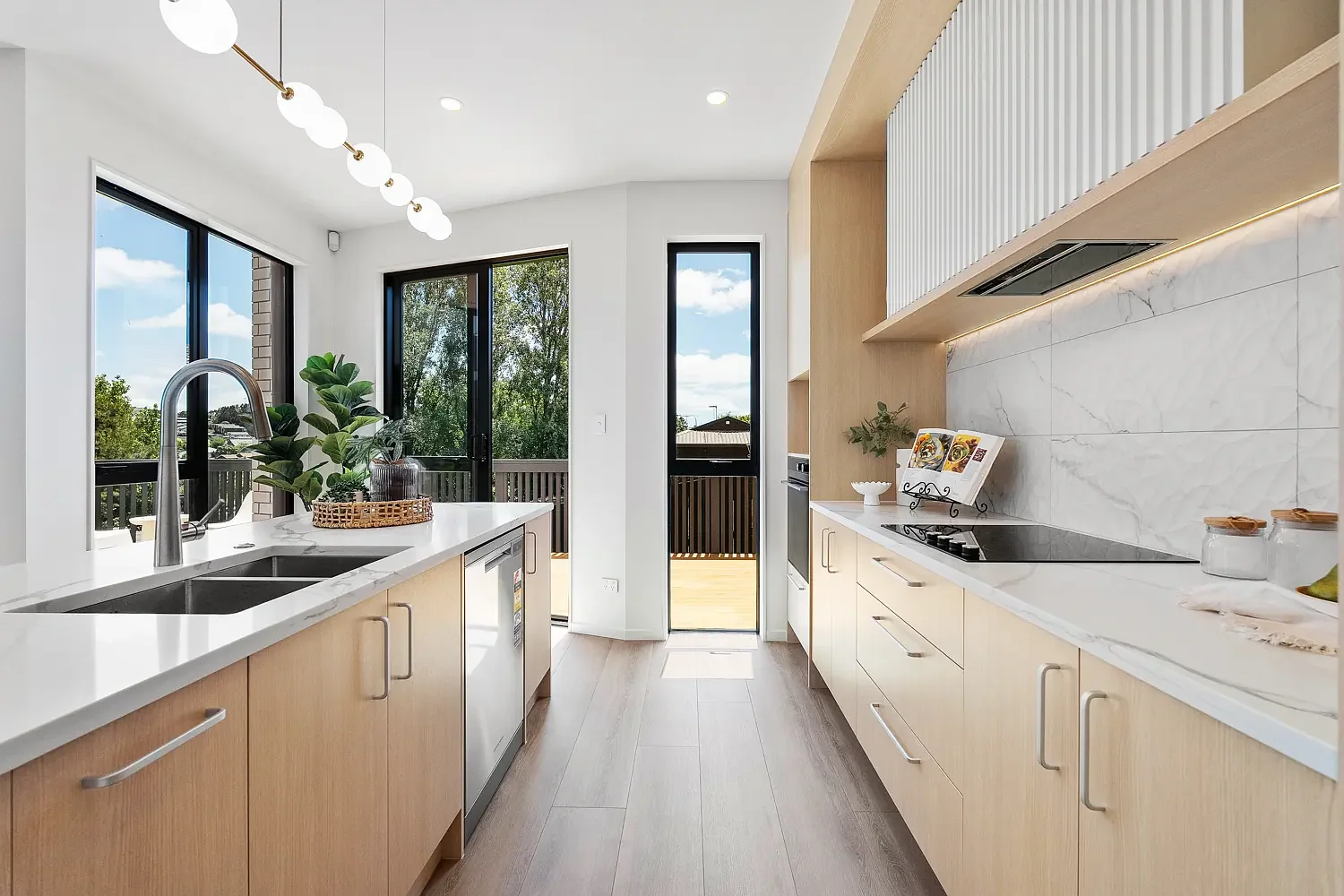 Modern kitchen with light wood cabinets, white marble countertops, a black cooktop, and large windows and door leading to a balcony with a view of trees and blue sky.