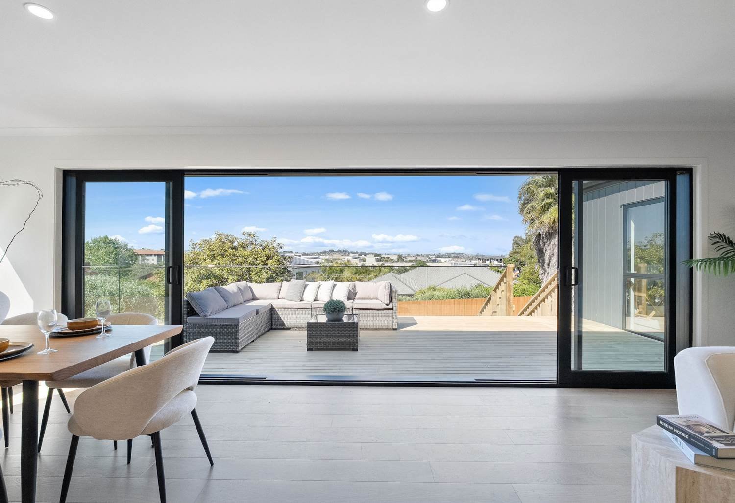 View of a spacious indoor living area opening onto a large outdoor deck with outdoor furniture and a view of trees, rooftops, and a blue sky with clouds.