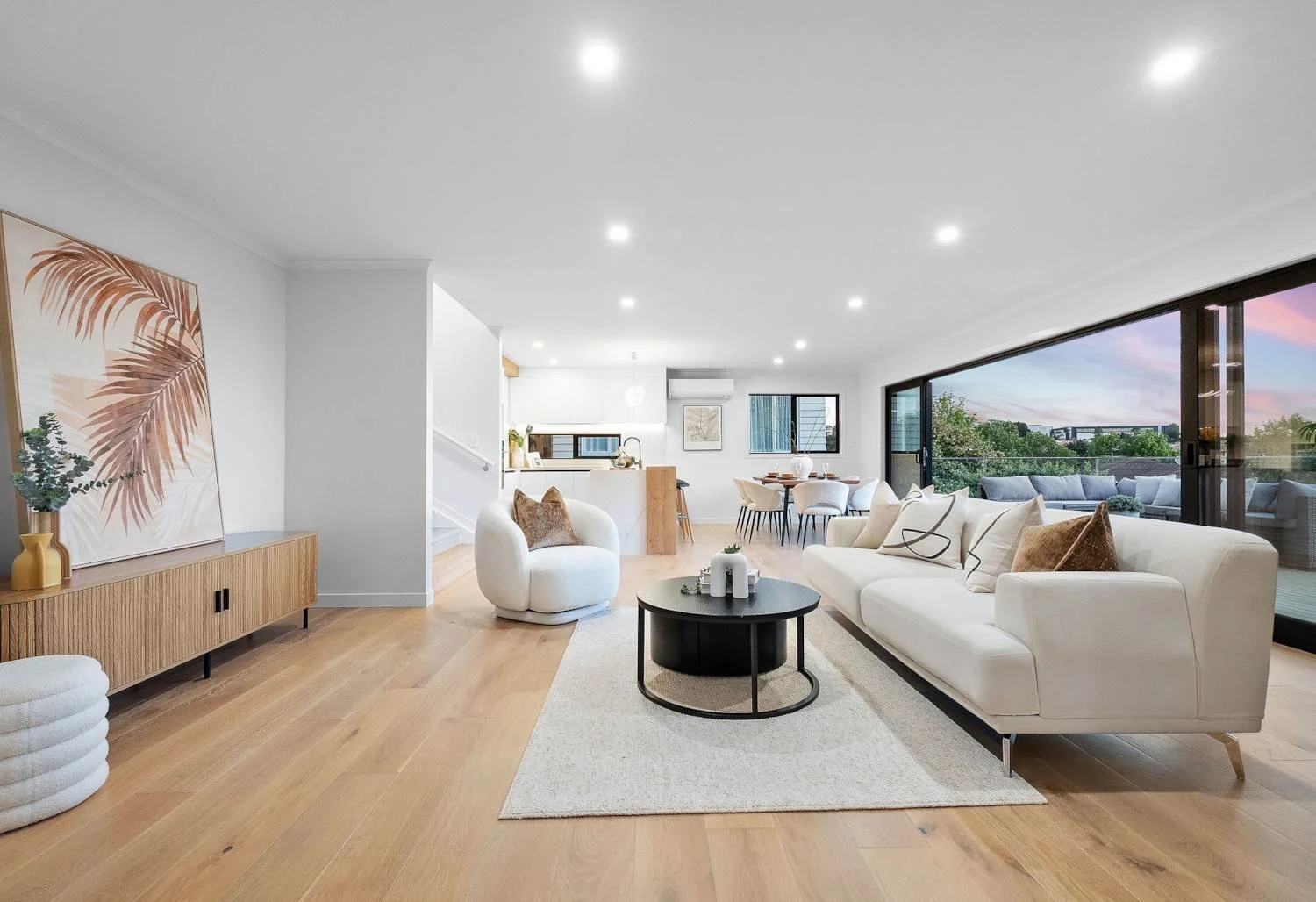 Modern open-concept living room with white sofa, round armchair, black coffee table, wooden sideboard with artwork, large sliding glass doors leading to balcony with outdoor seating, hardwood floors, and dining area in the background.