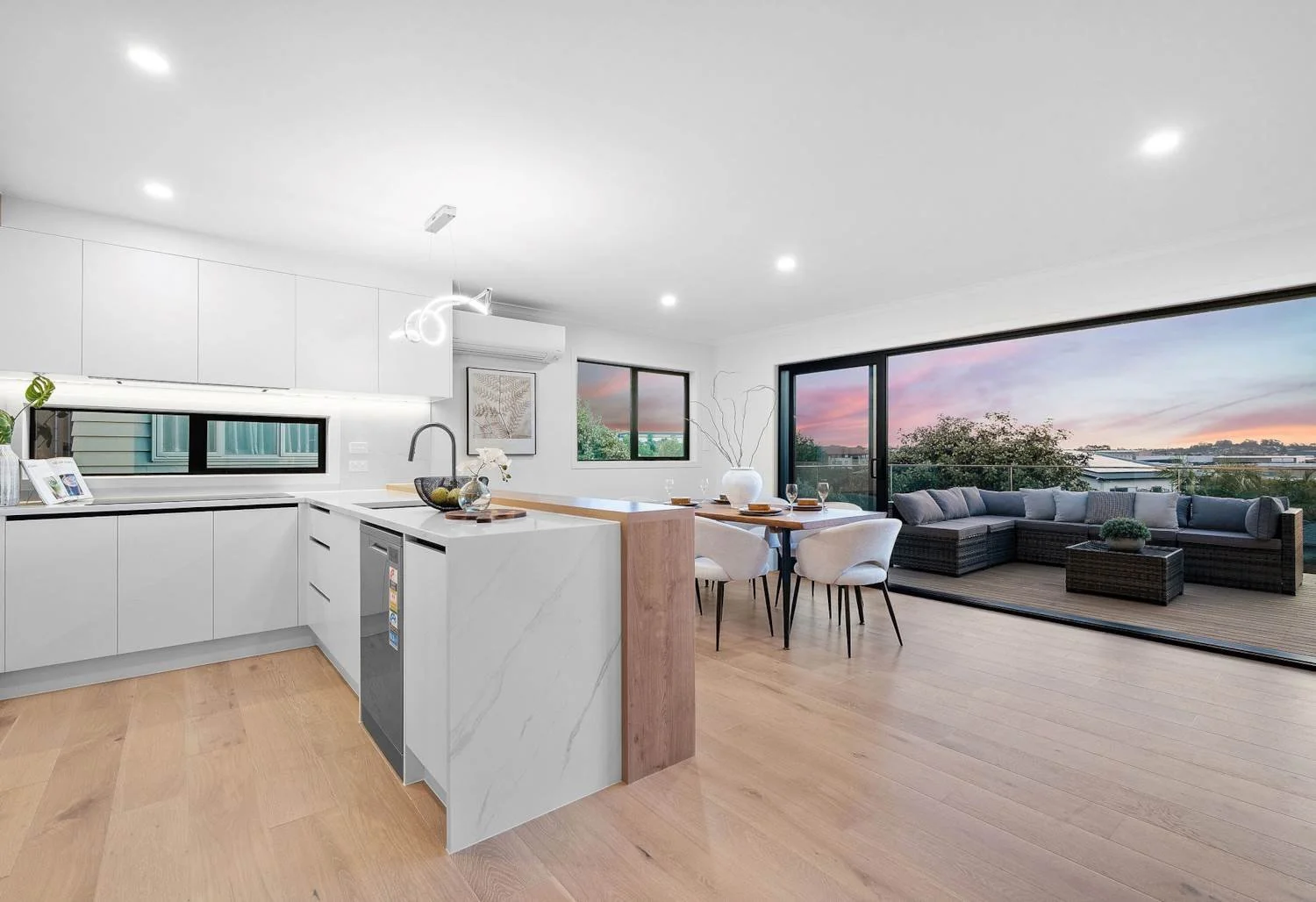 Modern, open-concept kitchen and living room with large sliding glass door opening to outdoor balcony with outdoor seating, sunset view, and light wood flooring.
