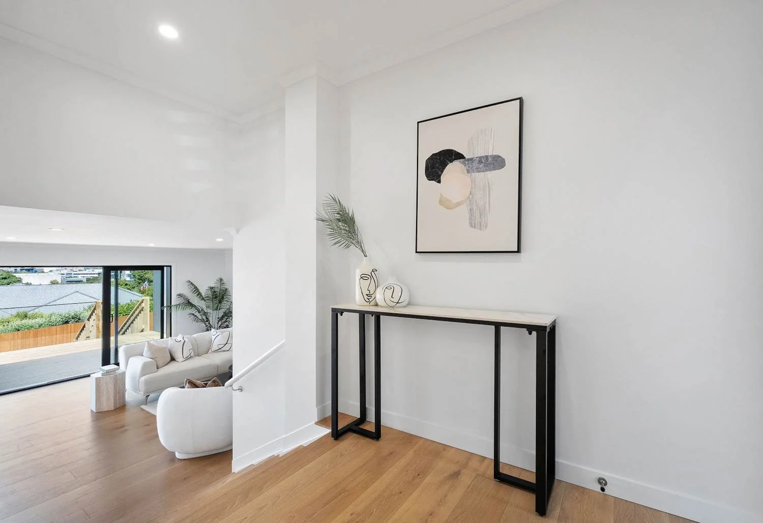 Modern living room interior with white walls, abstract art on the wall, a console table with decorative vases and a plant, and a view of a balcony with outdoor furniture and greenery