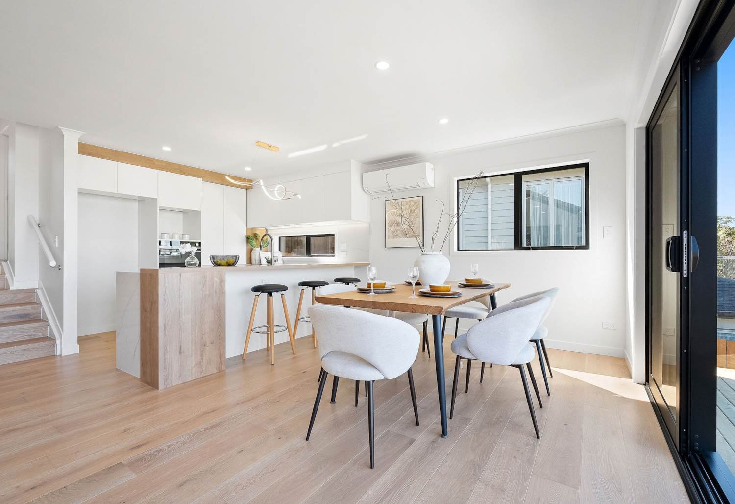 Open-concept kitchen and dining area with light wood flooring, white walls, modern furniture, and large glass sliding doors.