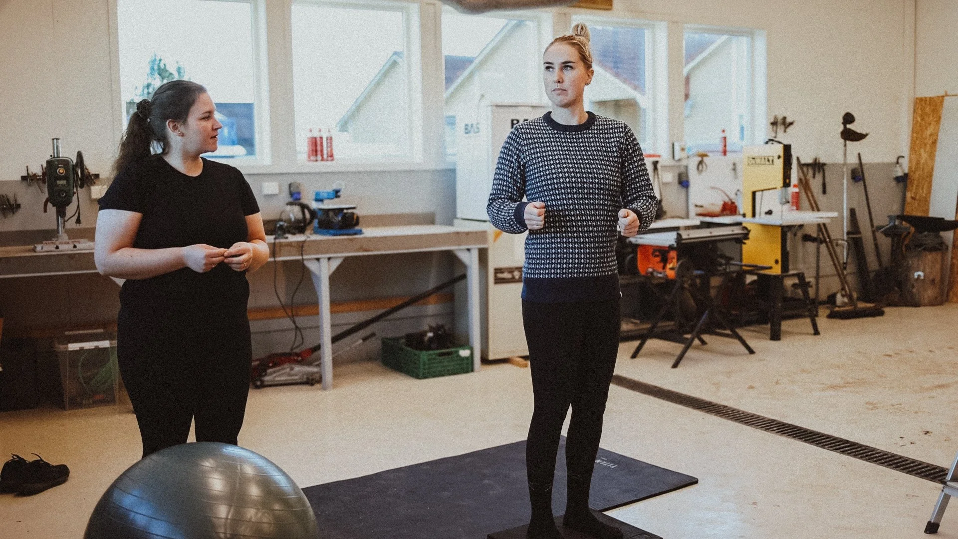 Two women standing in a fitness or rehabilitation space with workout equipment and tools, one of them is on a black mat with a fitness ball nearby.