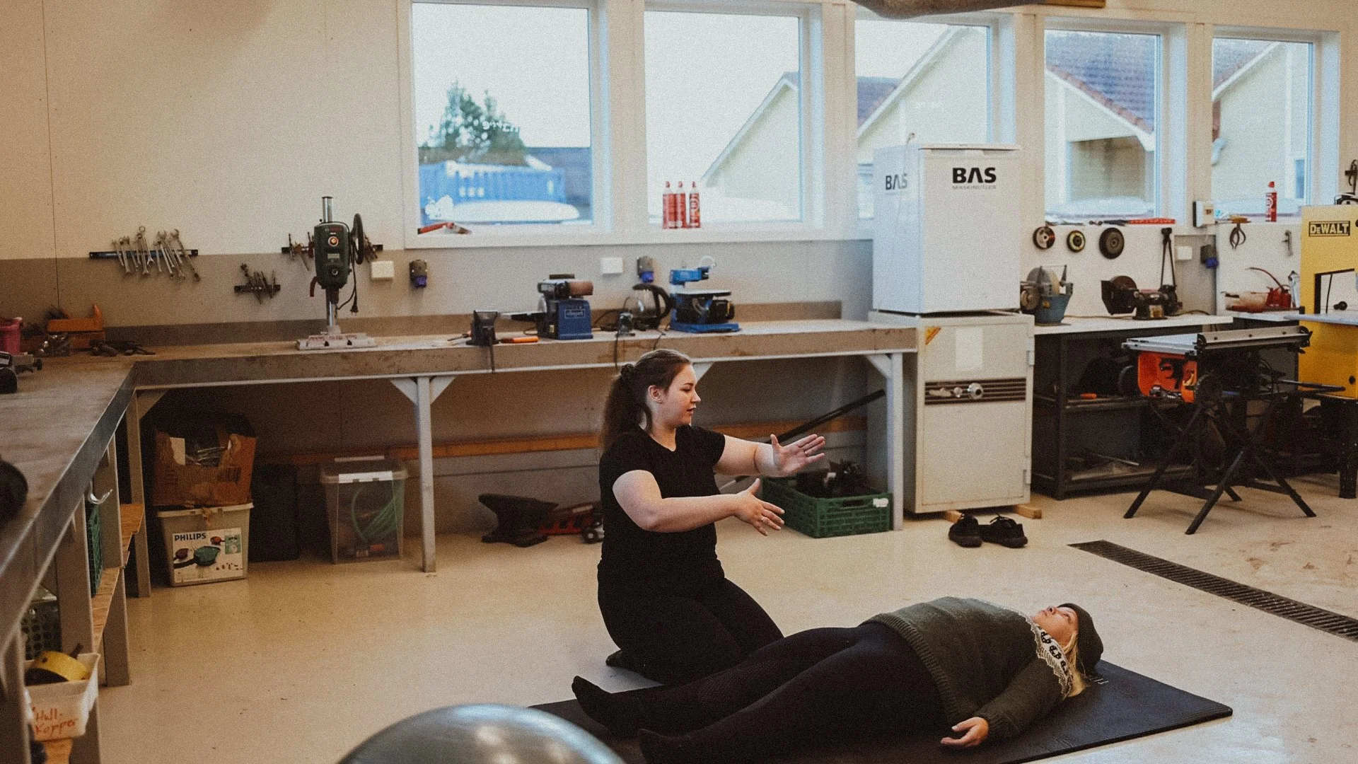 Two women practicing first aid or CPR on a woman lying on a black mat in a workshop or garage space.