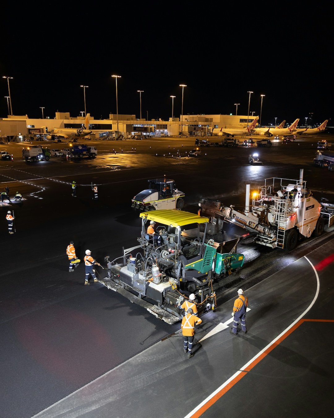 Nighttime scene at an airport runway showing workers paving or resurfacing the tarmac with heavy machinery. Visible airplanes are parked at gates in the background, with aircraft from Virgin airline. Airport terminal buildings and parking lot lights are also visible.