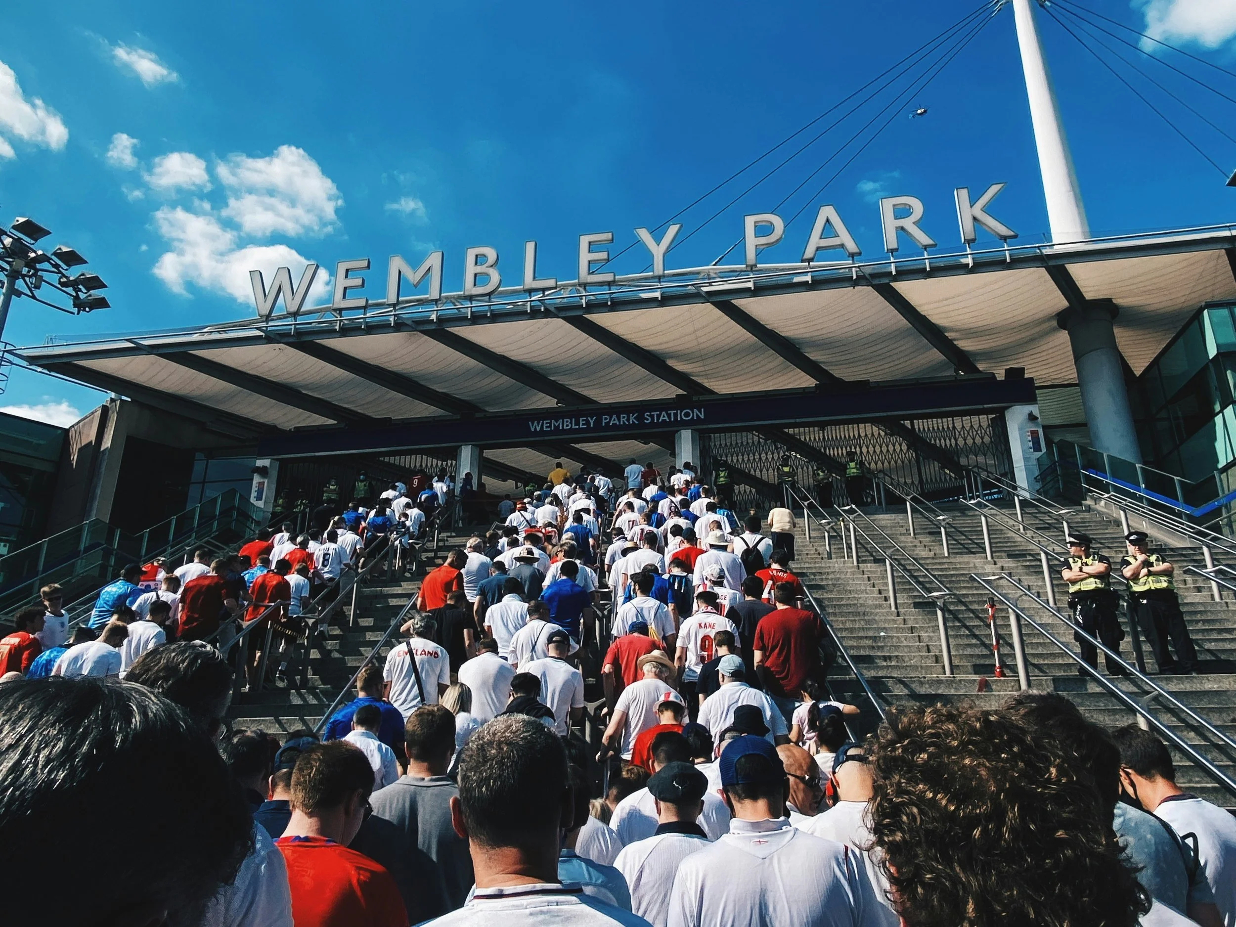 Crowd of people walking up the stairs to Wembley Park station under a clear blue sky with clouds, with the station sign visible.