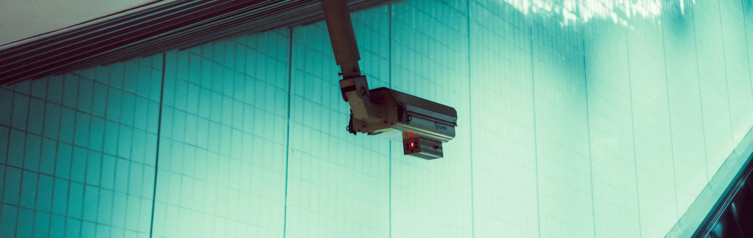 A security camera mounted on a wall, pointing downwards in a modern interior with glass wall panels.
