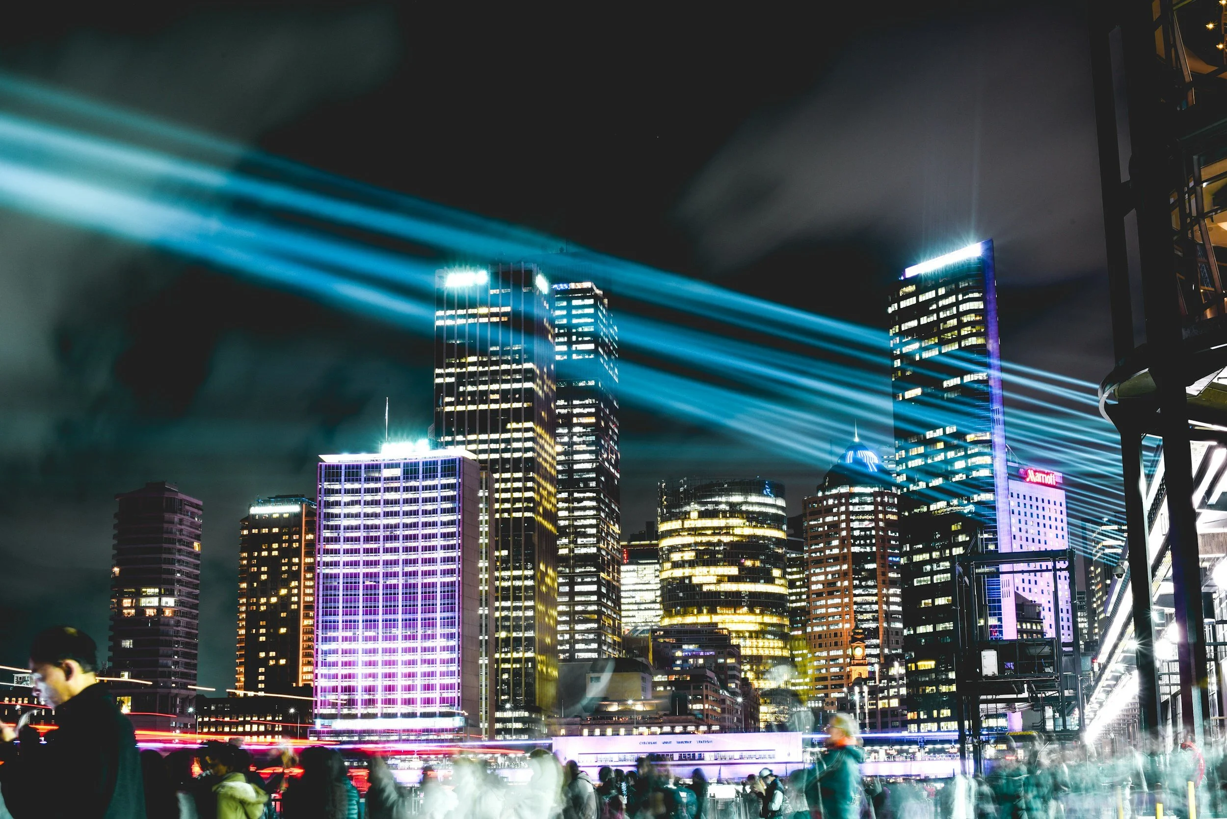Nighttime cityscape with tall buildings illuminated with colorful lights and beams of blue and purple light projected into the sky. Blurred people are visible in the foreground.