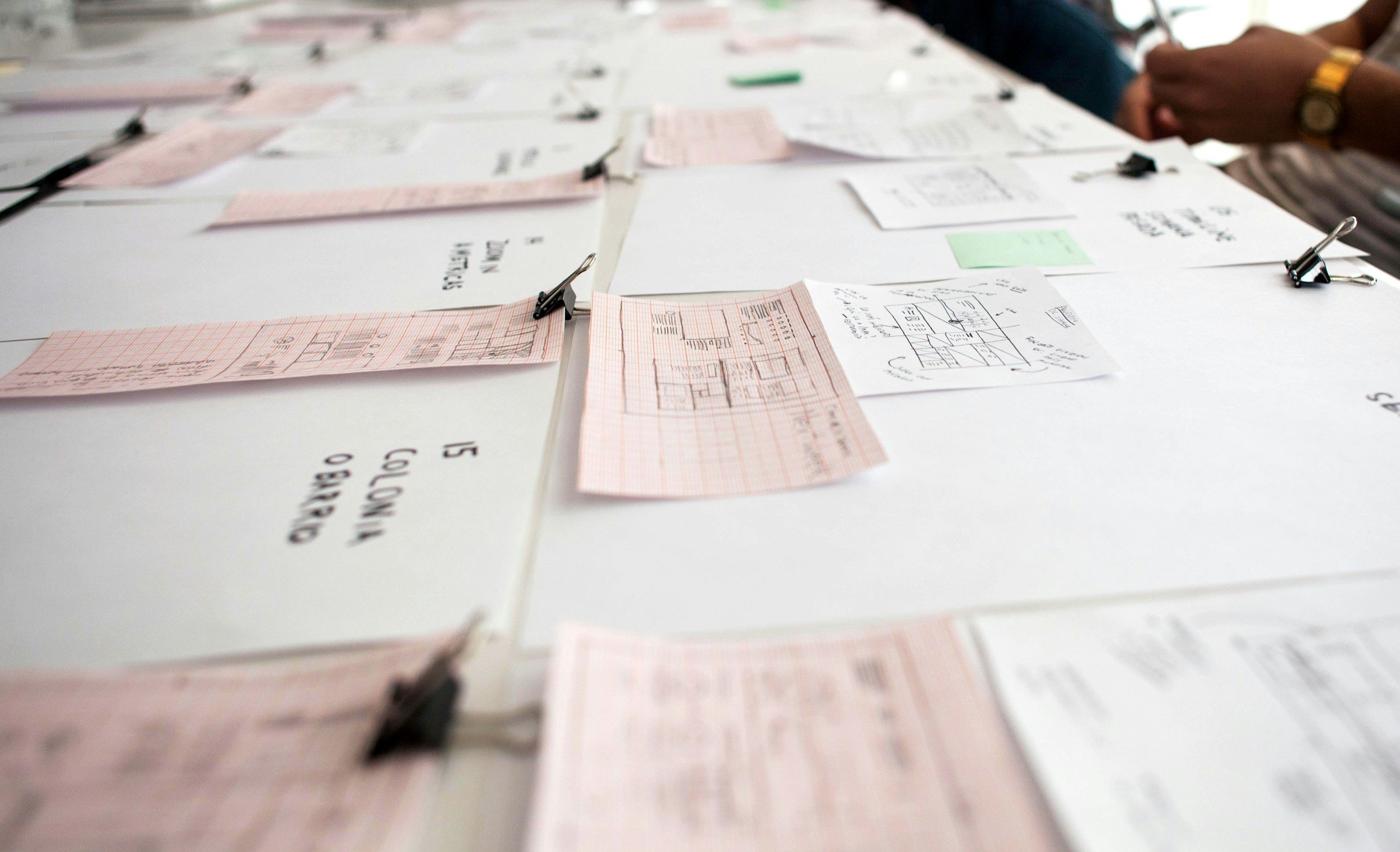 Several plan sheets and notes attached to a large white board or wall, with some people seen in the background.