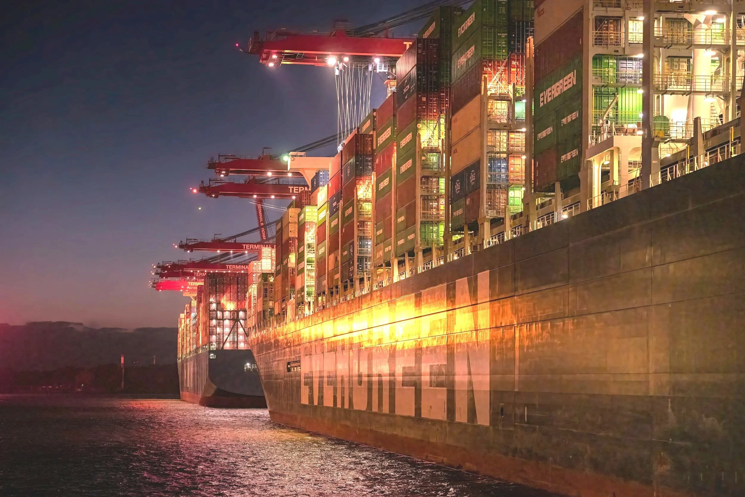 A large cargo ship docked at a port during night, with its numerous containers stacked and illuminated by port lights, and cranes visible in the background.