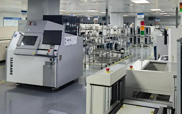 Inside an electronics manufacturing facility with automated machinery, conveyor belts, and equipment for assembling or inspecting electronic components.