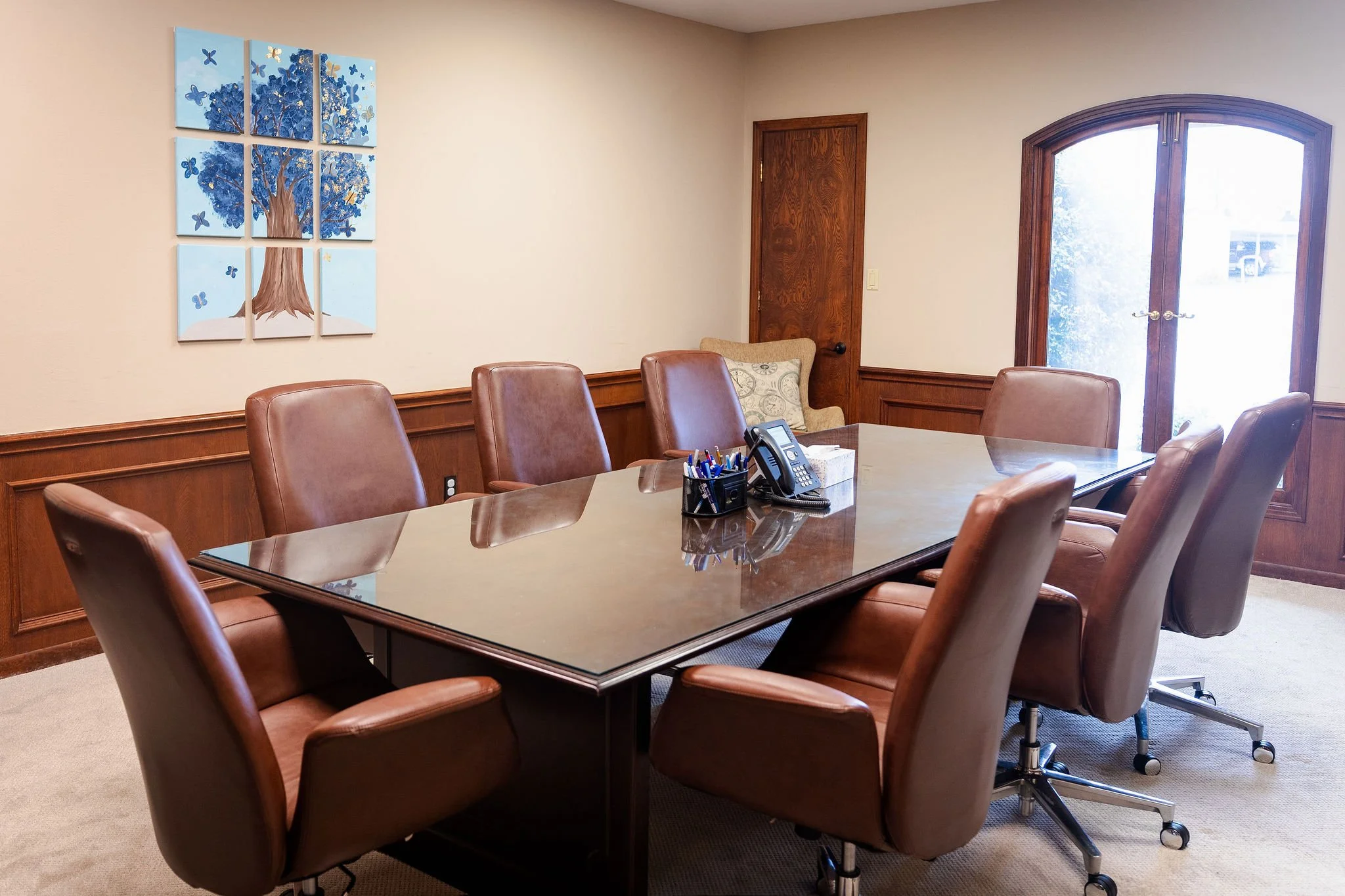 A conference room with a large wooden table surrounded by brown leather chairs. There is a black telephone and pen holder on the table. In the back, there are two windows with wooden frames and a beige armchair with patterned pillows. A multi-panel artwork depicting a blue tree with butterflies hangs on the beige wall.