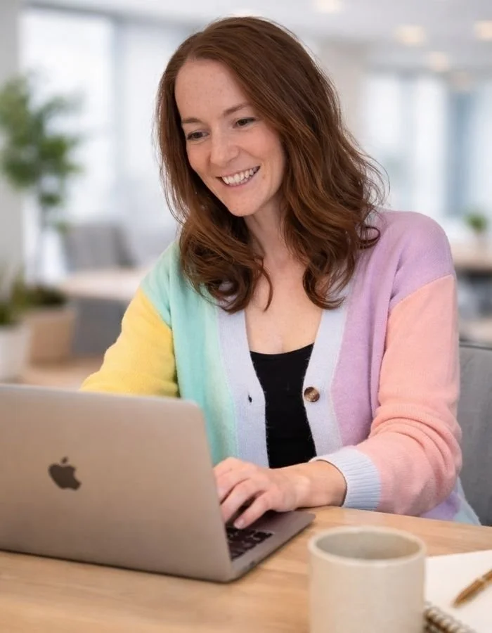 A woman with red hair smiling while working on a silver MacBook in an office setting.