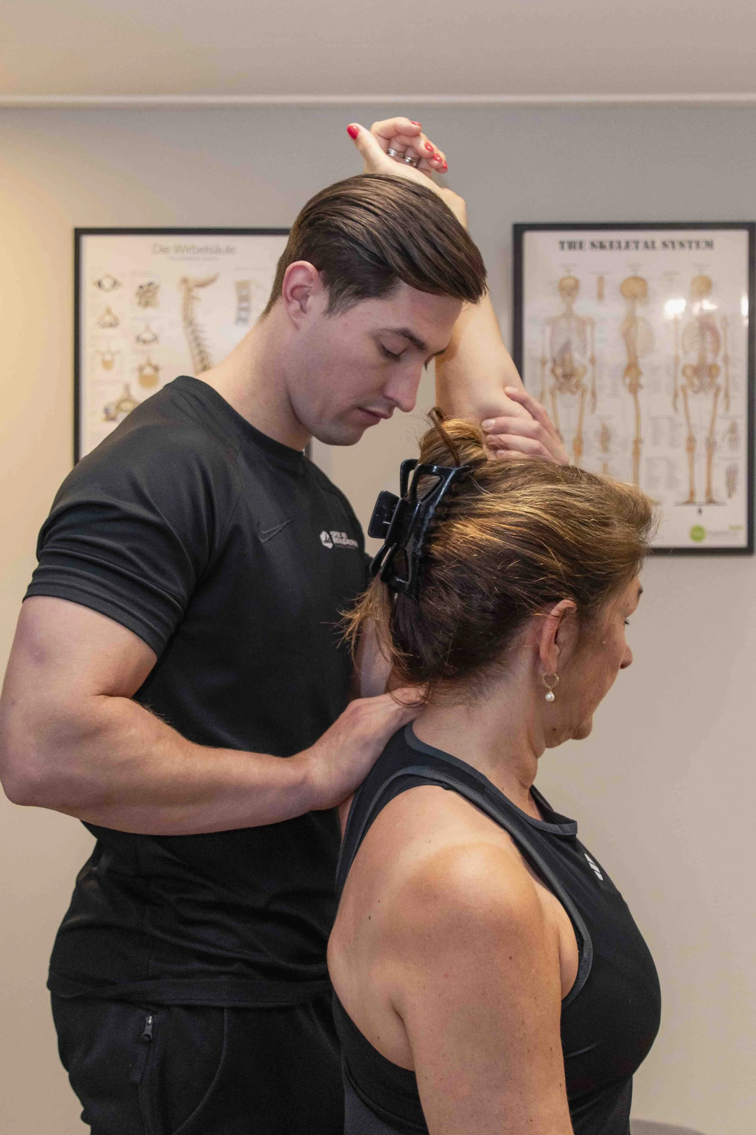 Een man geeft een massage of therapie aan een vrouw die uitrust op een massagetafel. Ze dragen beide zwarte kleding en de achtergrond toont anatomische posters.