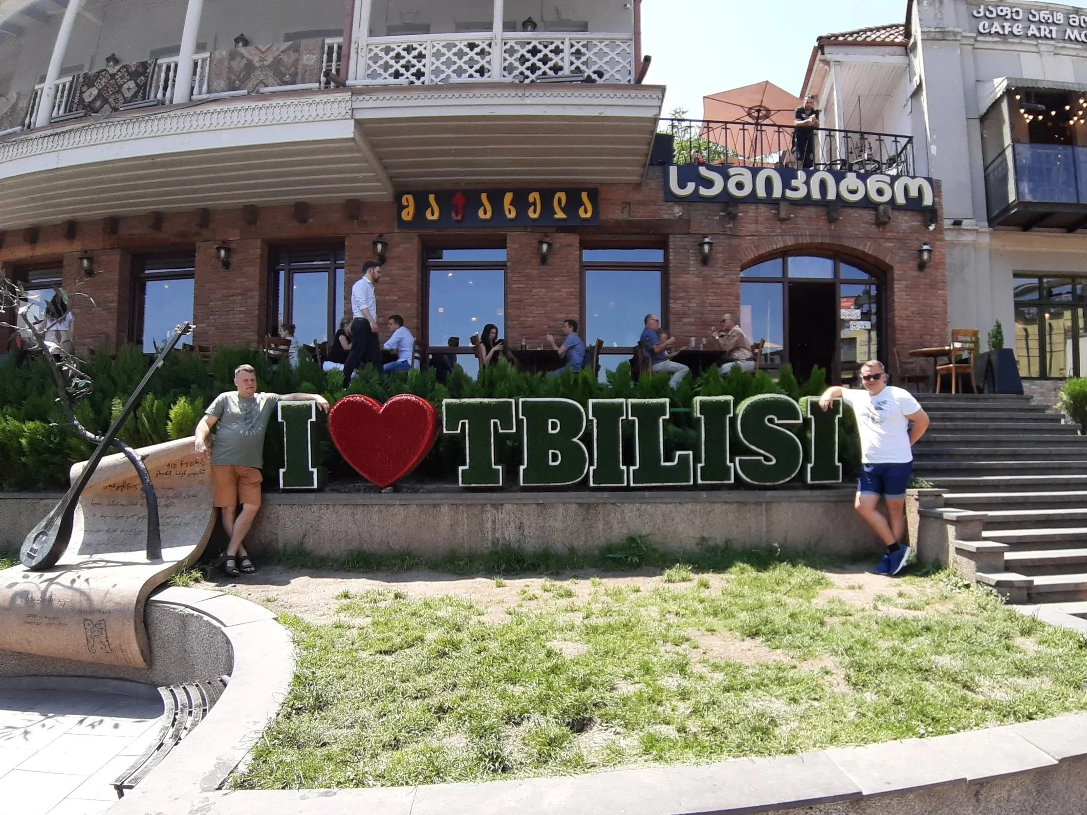 Three people standing in front of a sign that says "I ♥ Tbilisi" in front of a brick building with outdoor seating and large windows, with people dining inside, on a sunny day.
