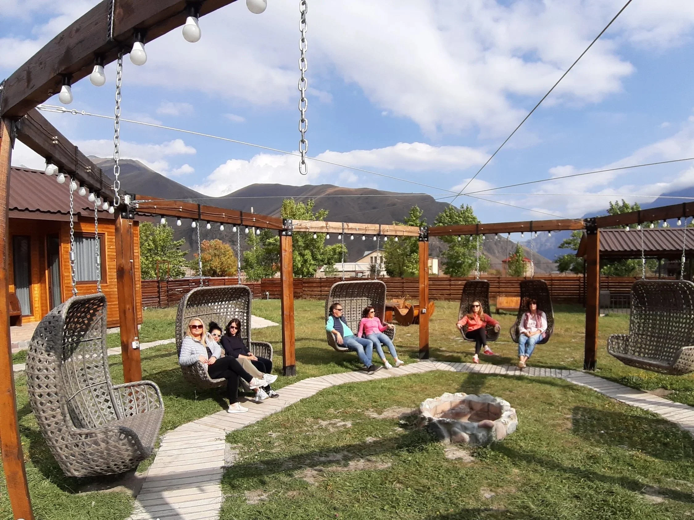 Six people relaxing in outdoor hanging chairs in a grassy backyard with mountains in the background. The sky is partly cloudy and a fire pit is in the center.