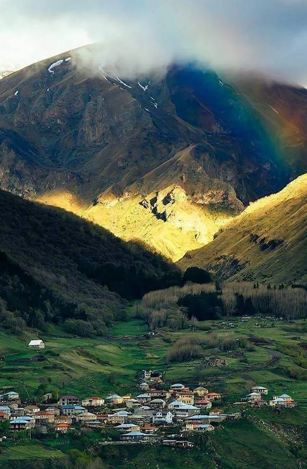 Scenic view of a mountain landscape with a small village at the base, lush green hills, and a rainbow on the right side.