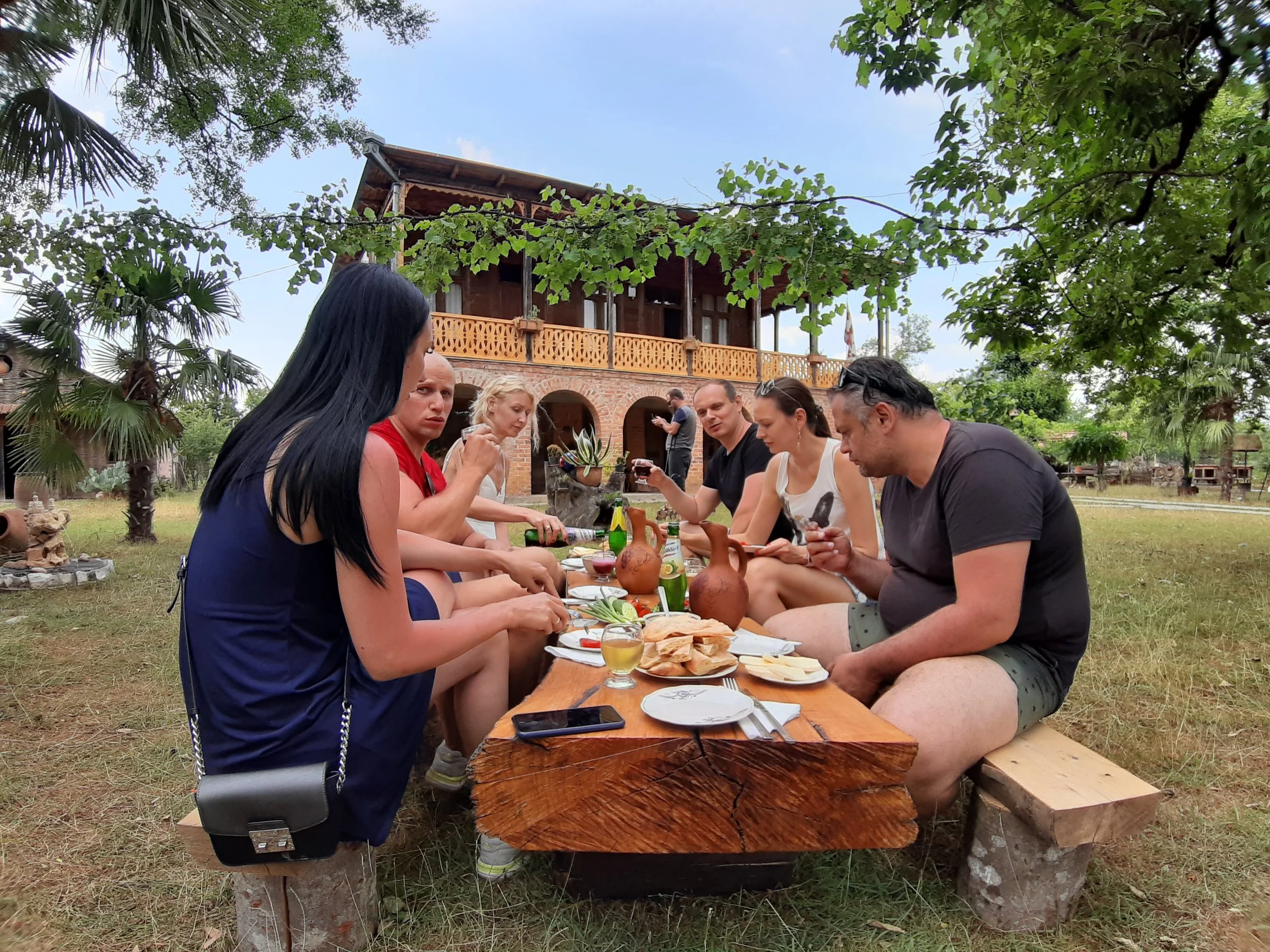 People having an outdoor meal at a wooden table in a garden with a large brick house in the background, surrounded by trees and greenery.