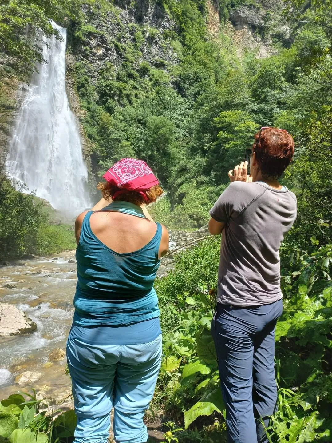Two women are standing near a waterfall surrounded by green trees and rocks, one is taking a photo with her phone.