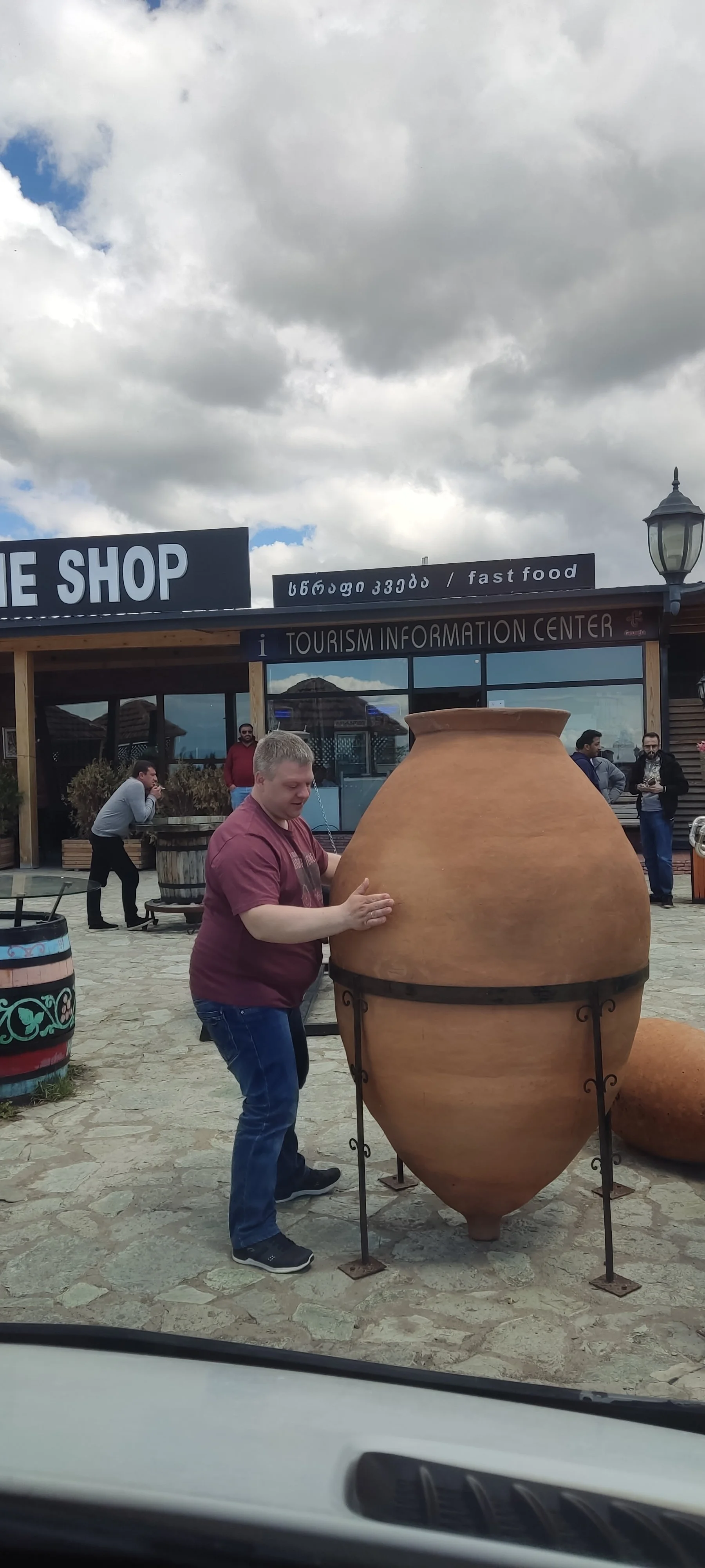 A man touching a large ancient-looking clay pot outdoors in front of a tourism information center building.